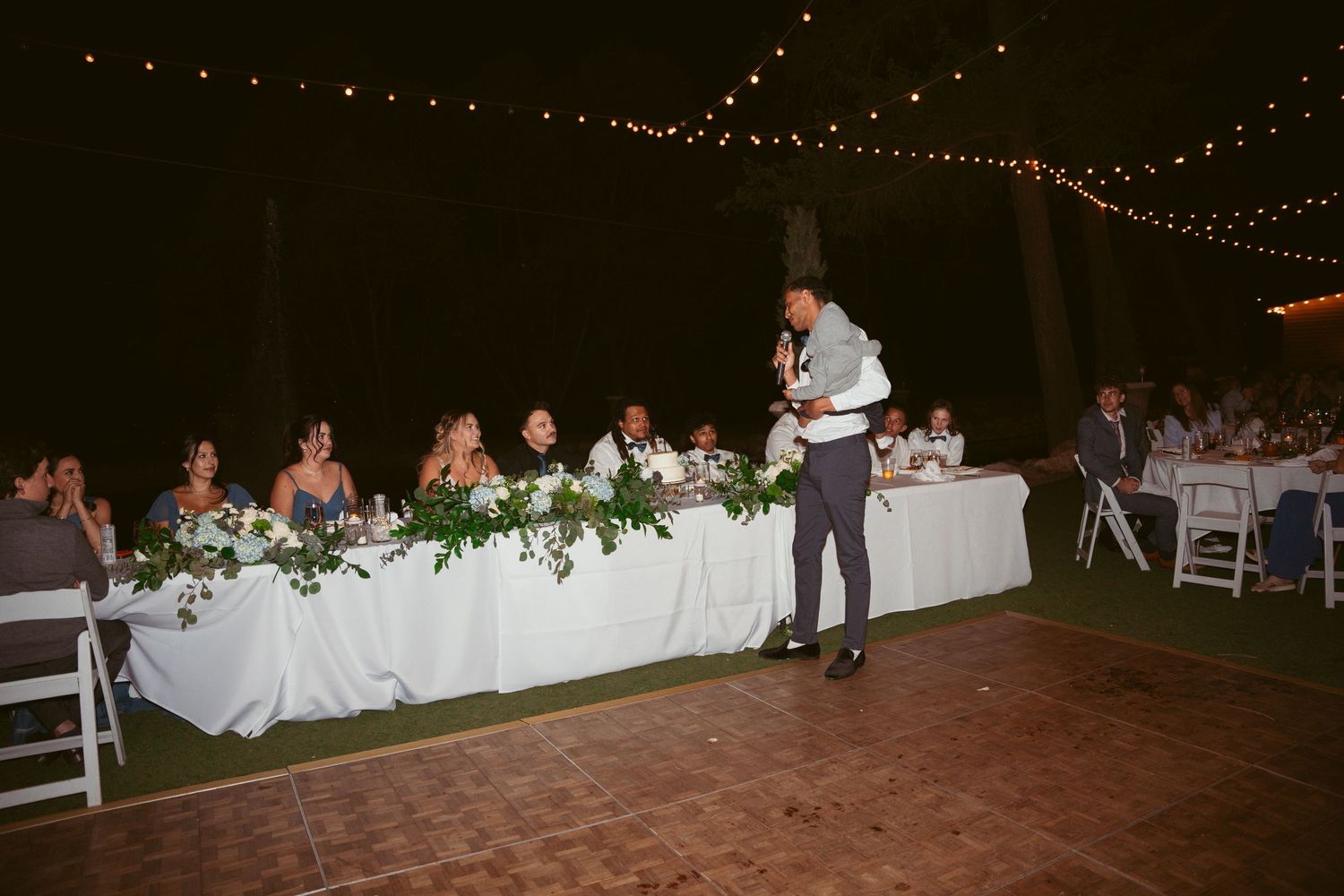 A speaker addresses guests at a wedding reception with string lights overhead and a decorated head table.