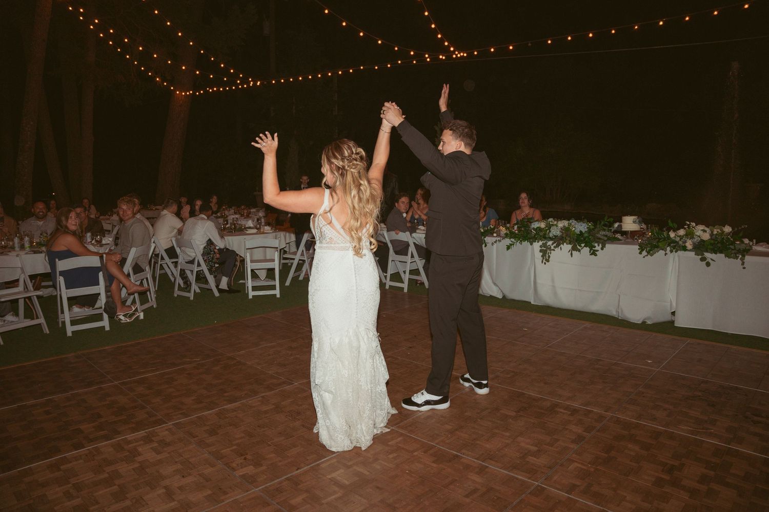 Wedding party dancing on a wooden dance floor under twinkling string lights at dusk.