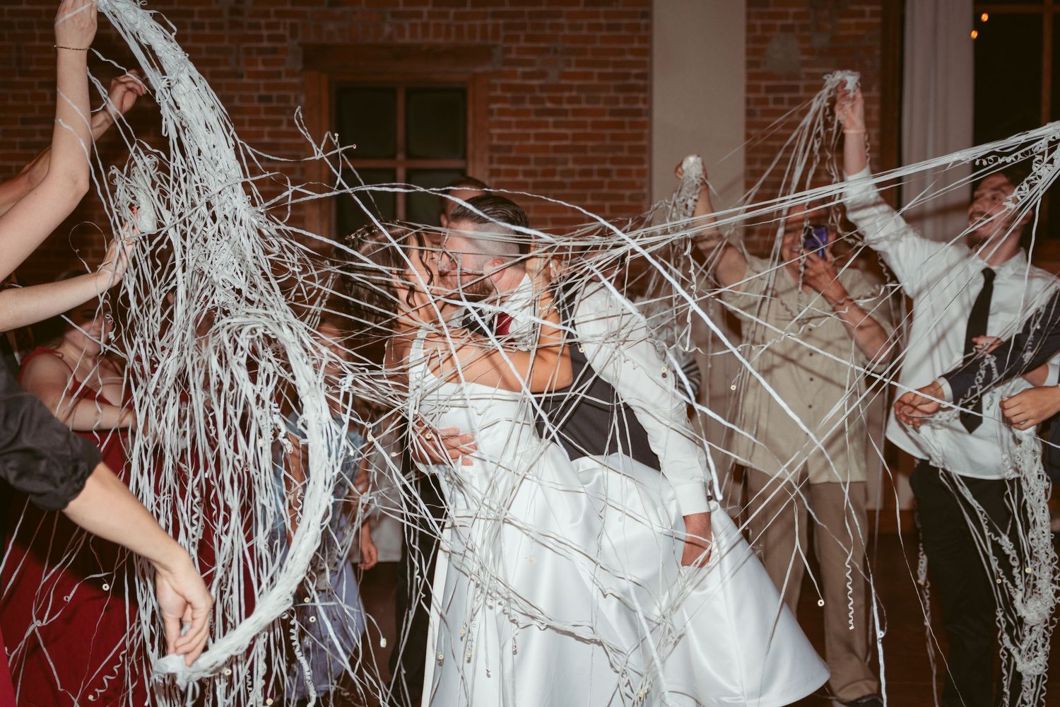 Wedding party guests toss white streamers in celebration at indoor brick venue with string lights.