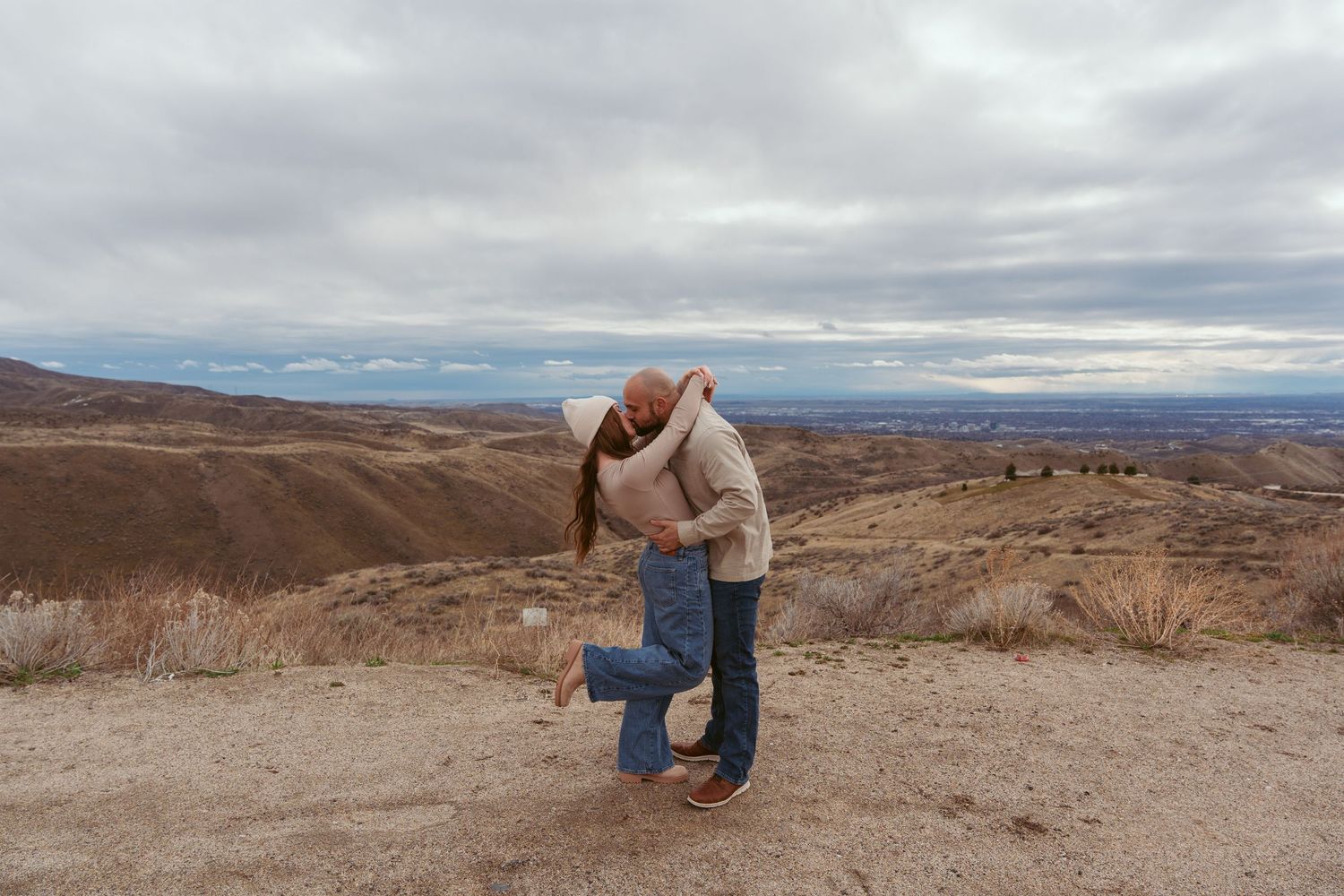 A couple embraces on a hilltop overlooking rolling brown hills and mountains under cloudy skies.