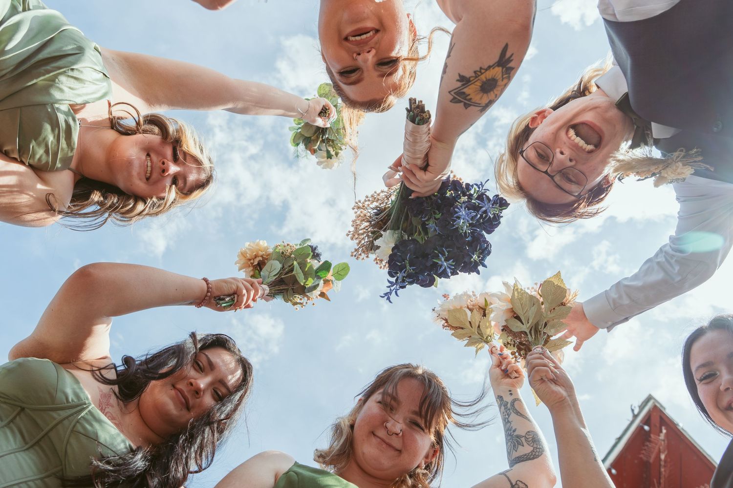 Wedding party forms circle looking down at camera against blue sky.
