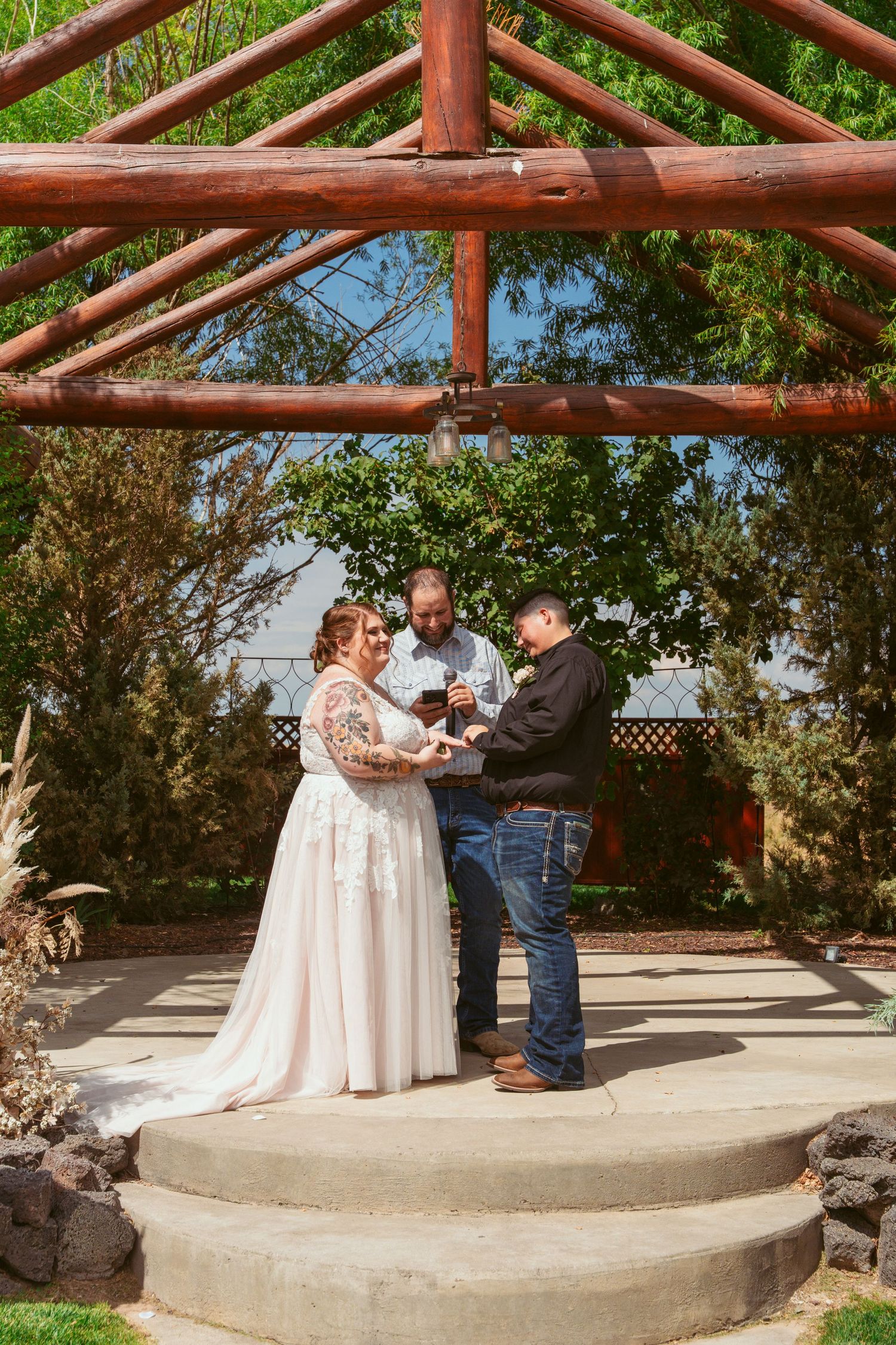 A wedding ceremony takes place under a rustic wooden pergola on a raised concrete platform in an outdoor garden setting.
