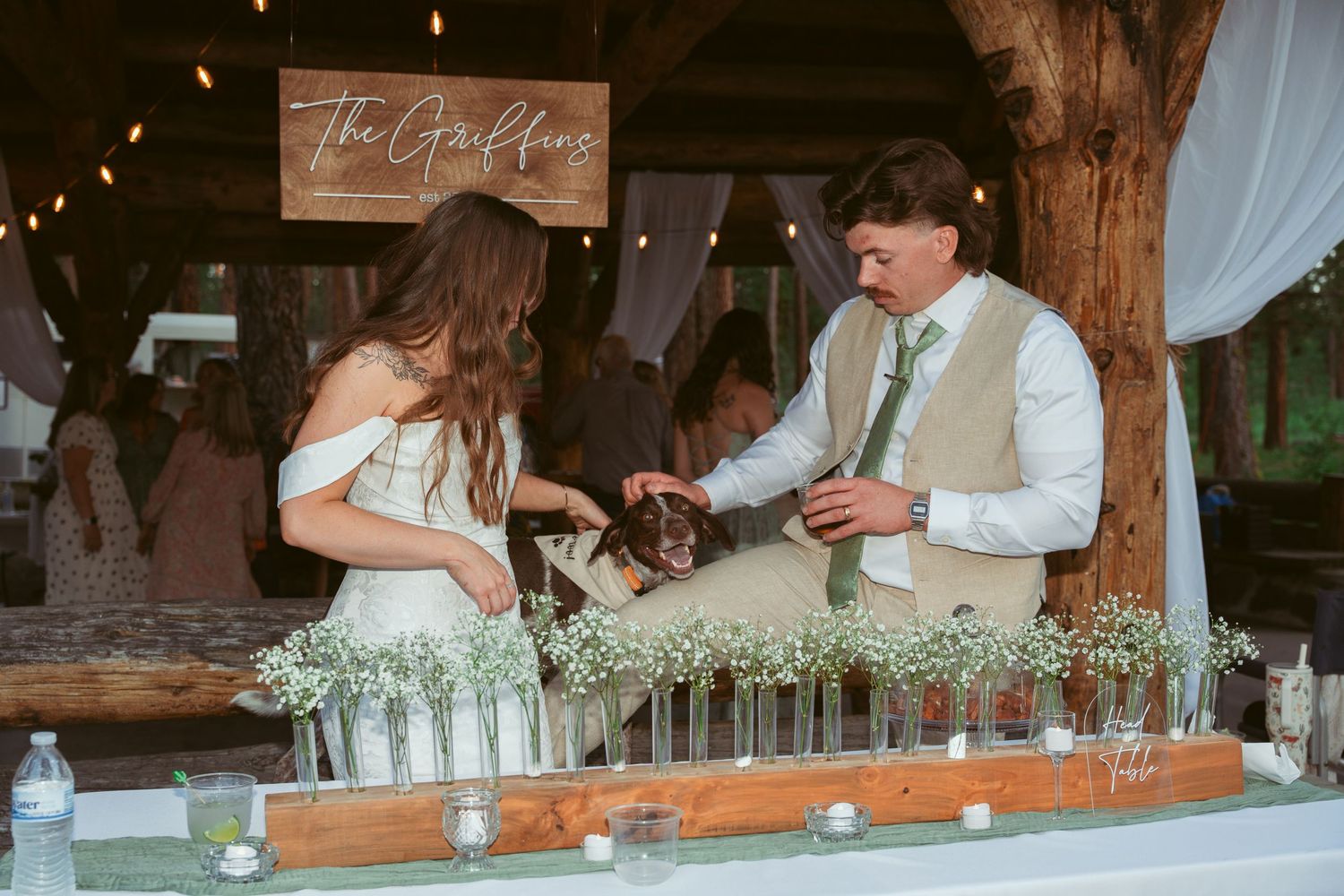 Wedding reception bar setup in rustic barn venue with string lights and wooden beams.