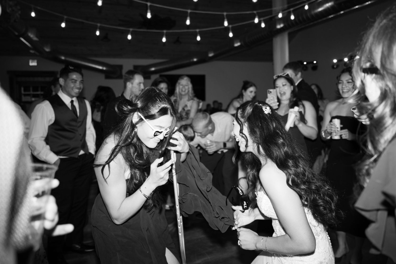 Wedding reception guests dancing and celebrating on a dimly lit dance floor with string lights overhead.
