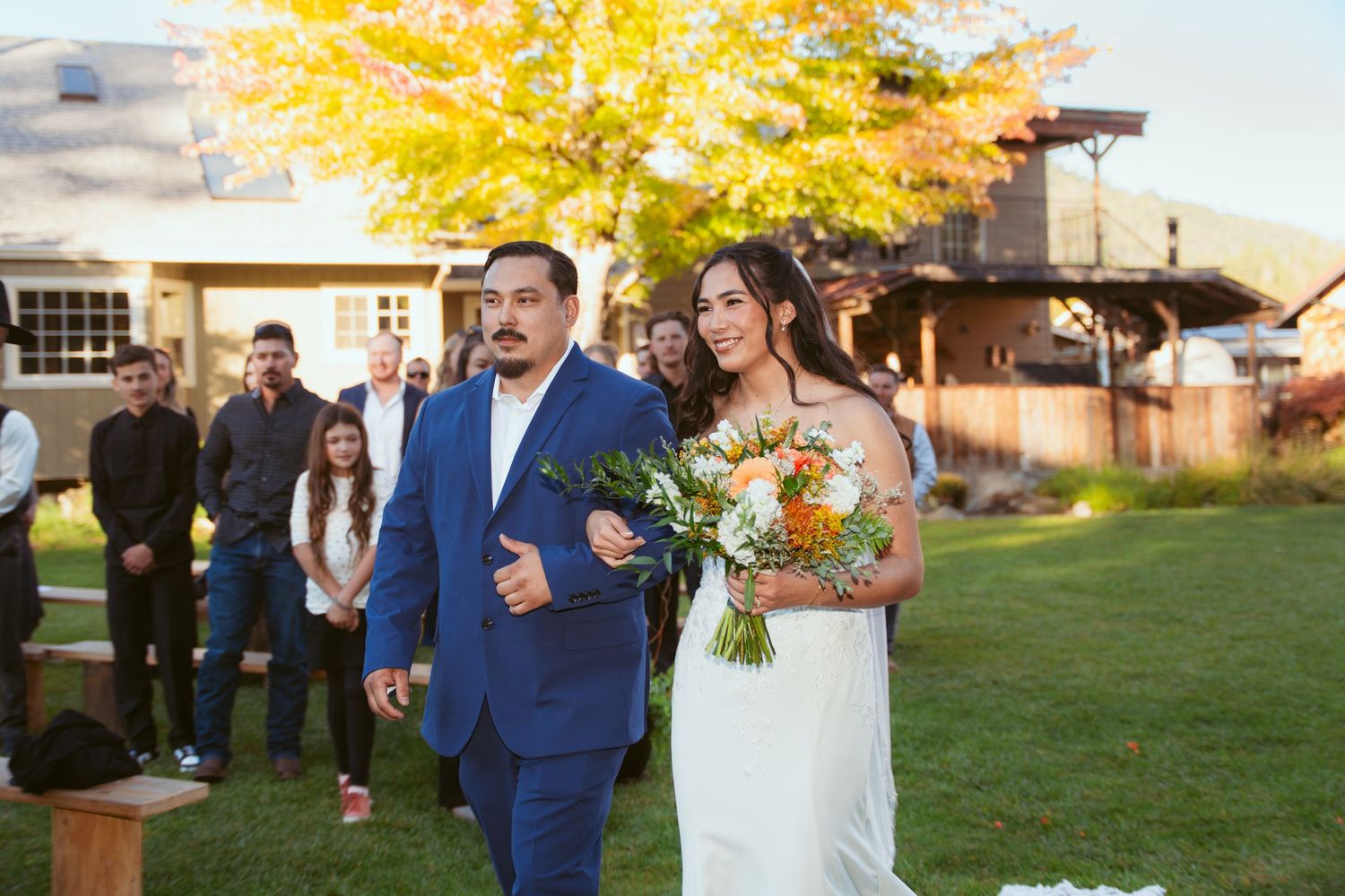Wedding ceremony processional on grass with autumn foliage and barn setting.
