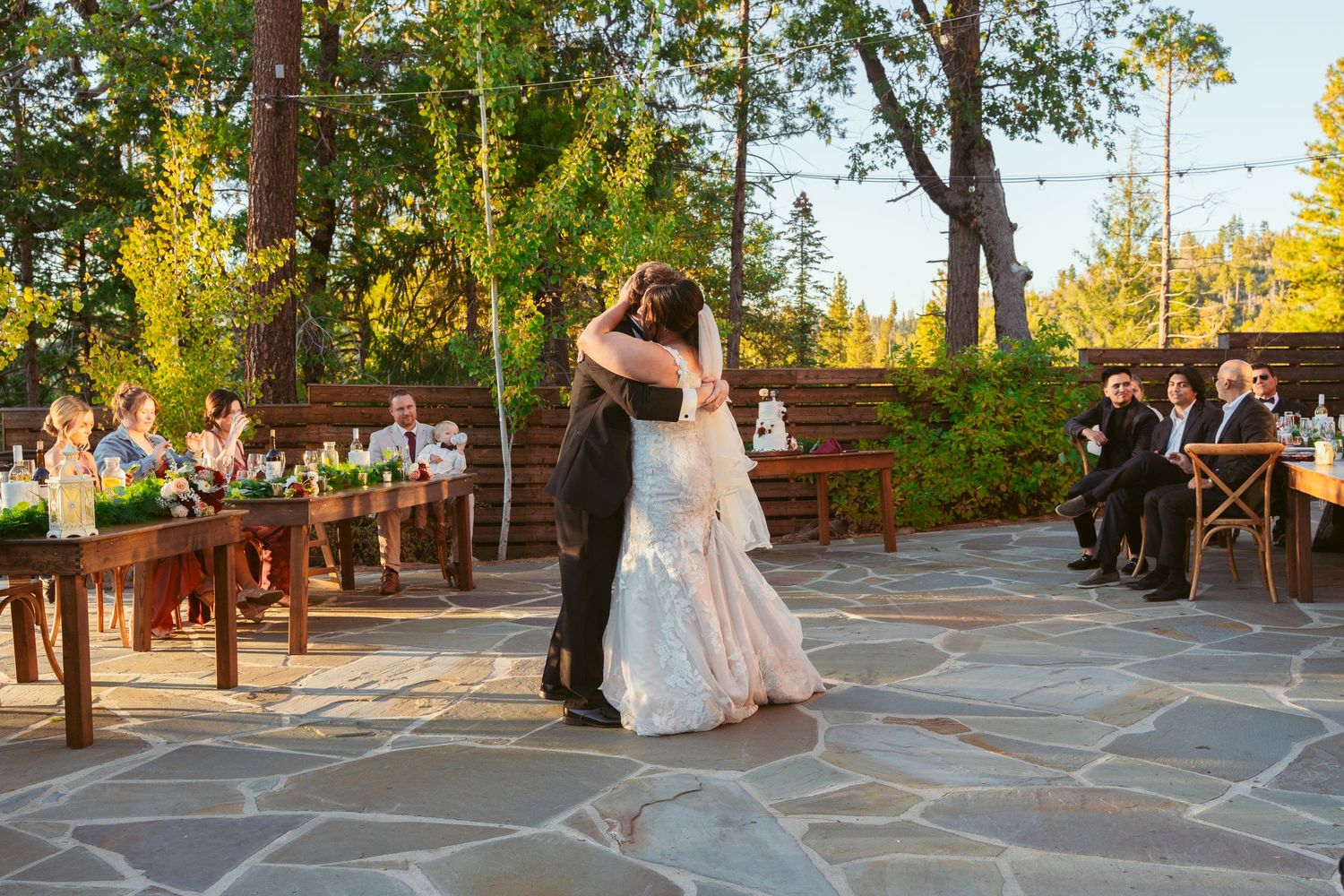 Wedding celebration on outdoor stone patio with guests watching couple's first dance at sunset.