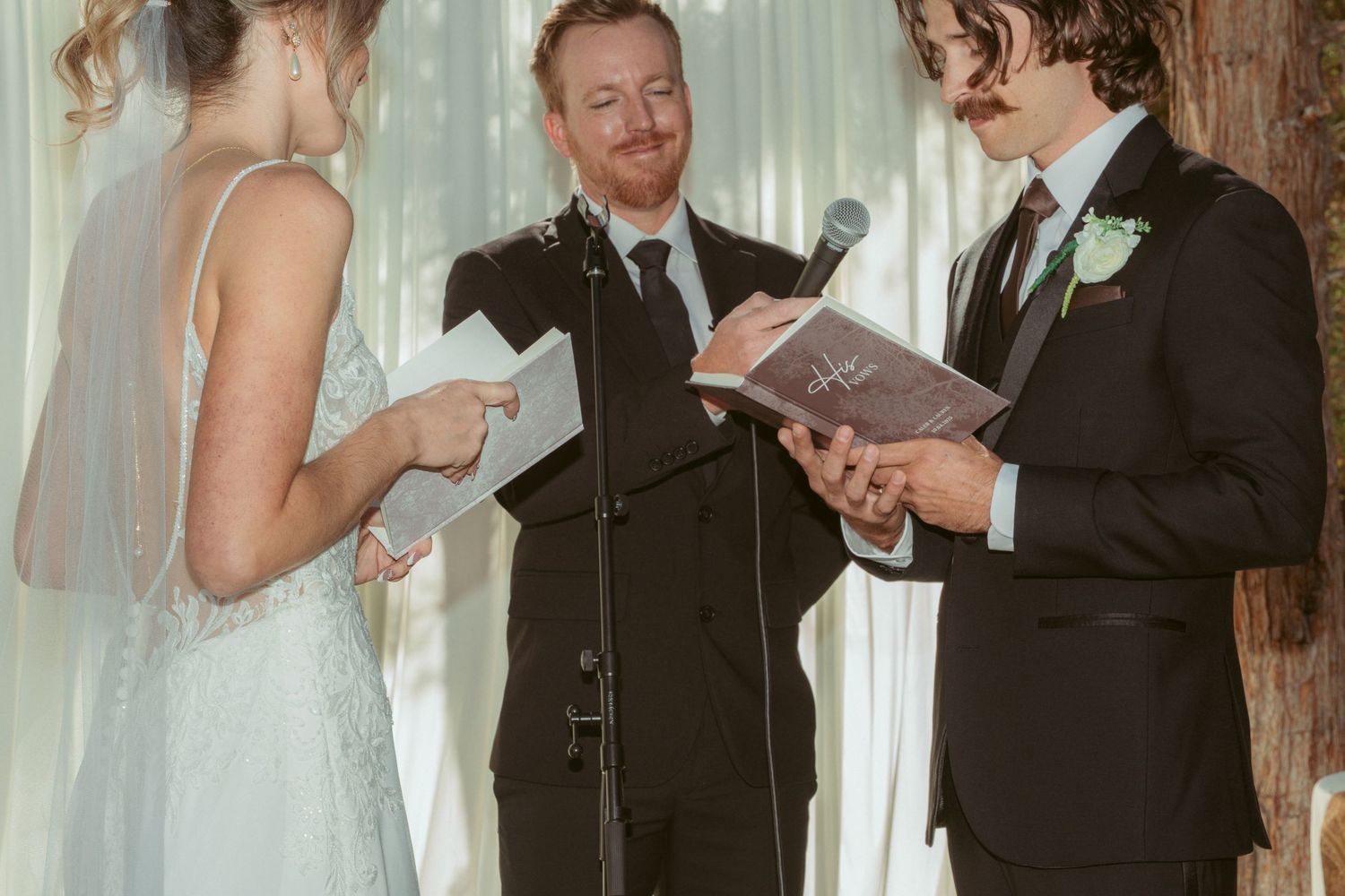 Wedding ceremony participants exchanging vows in front of sheer white curtains outdoors.
