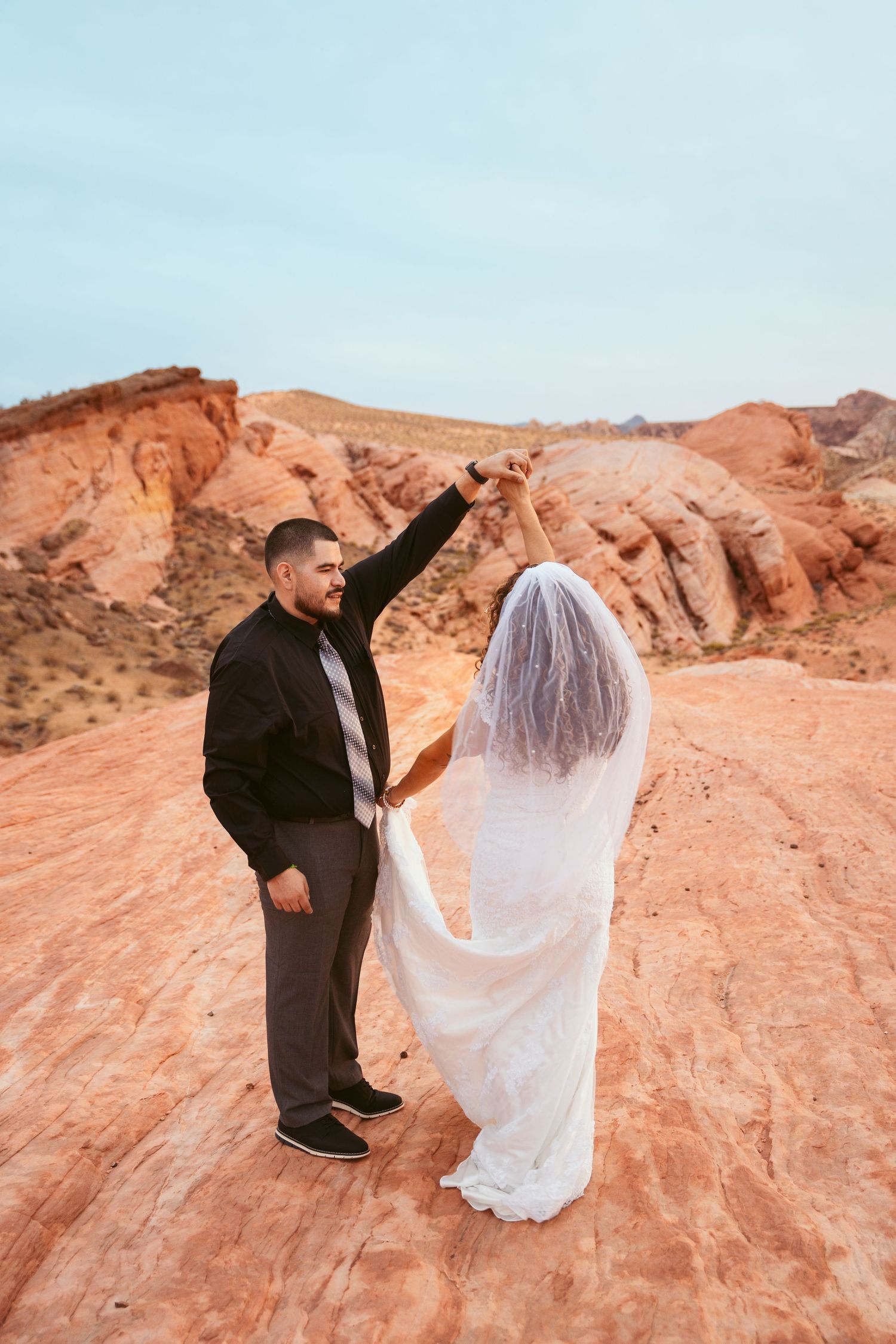 A couple in wedding attire poses against dramatic red rock formations in a desert landscape.