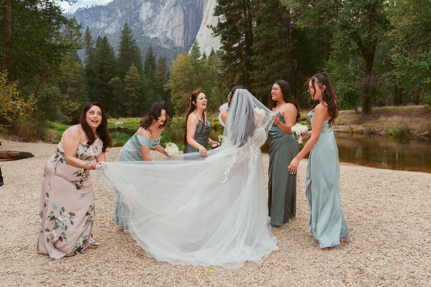 Wedding party arranges bride's long train on rocky beach with mountain backdrop.