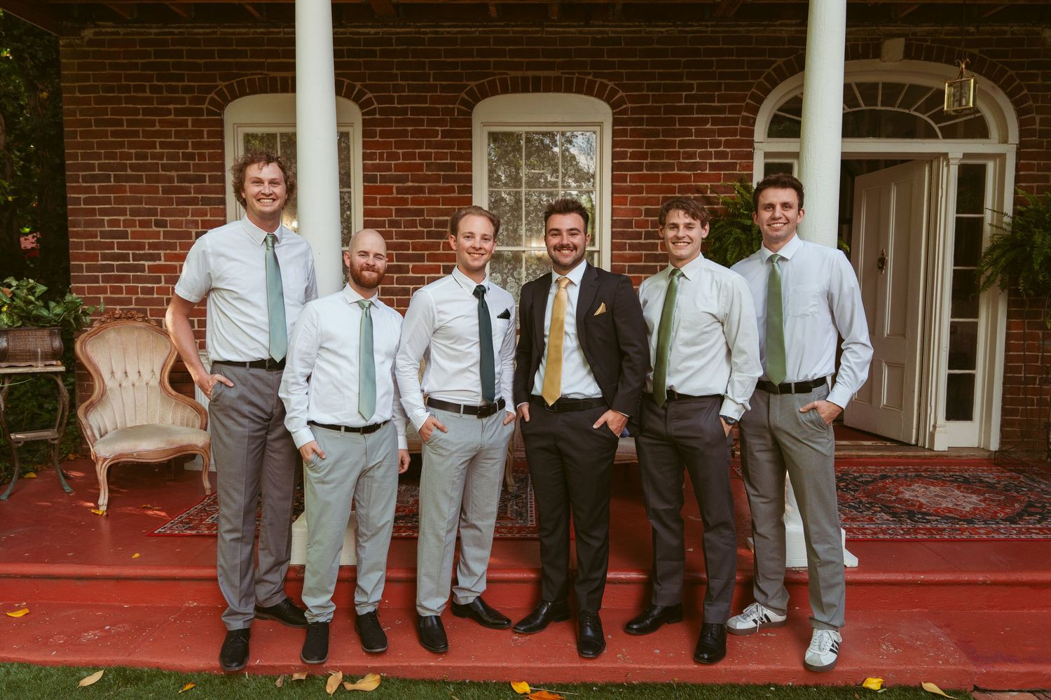 Groomsmen in light gray suits pose playfully on brick porch with white columns.