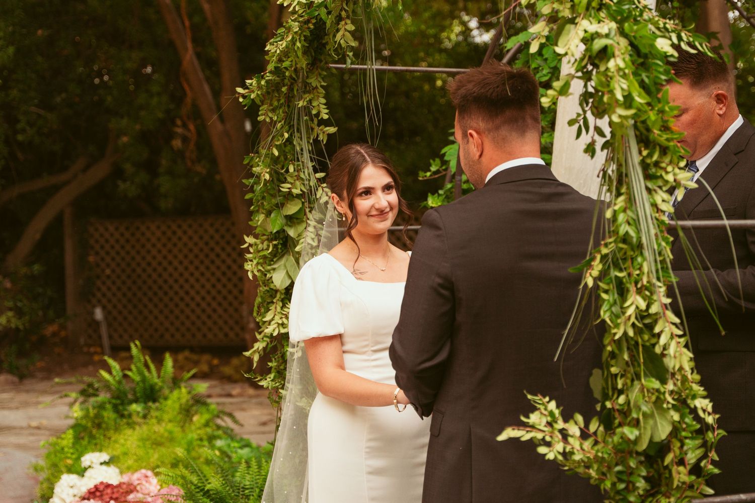 A couple exchanges vows beneath a greenery-adorned arch during their garden wedding.