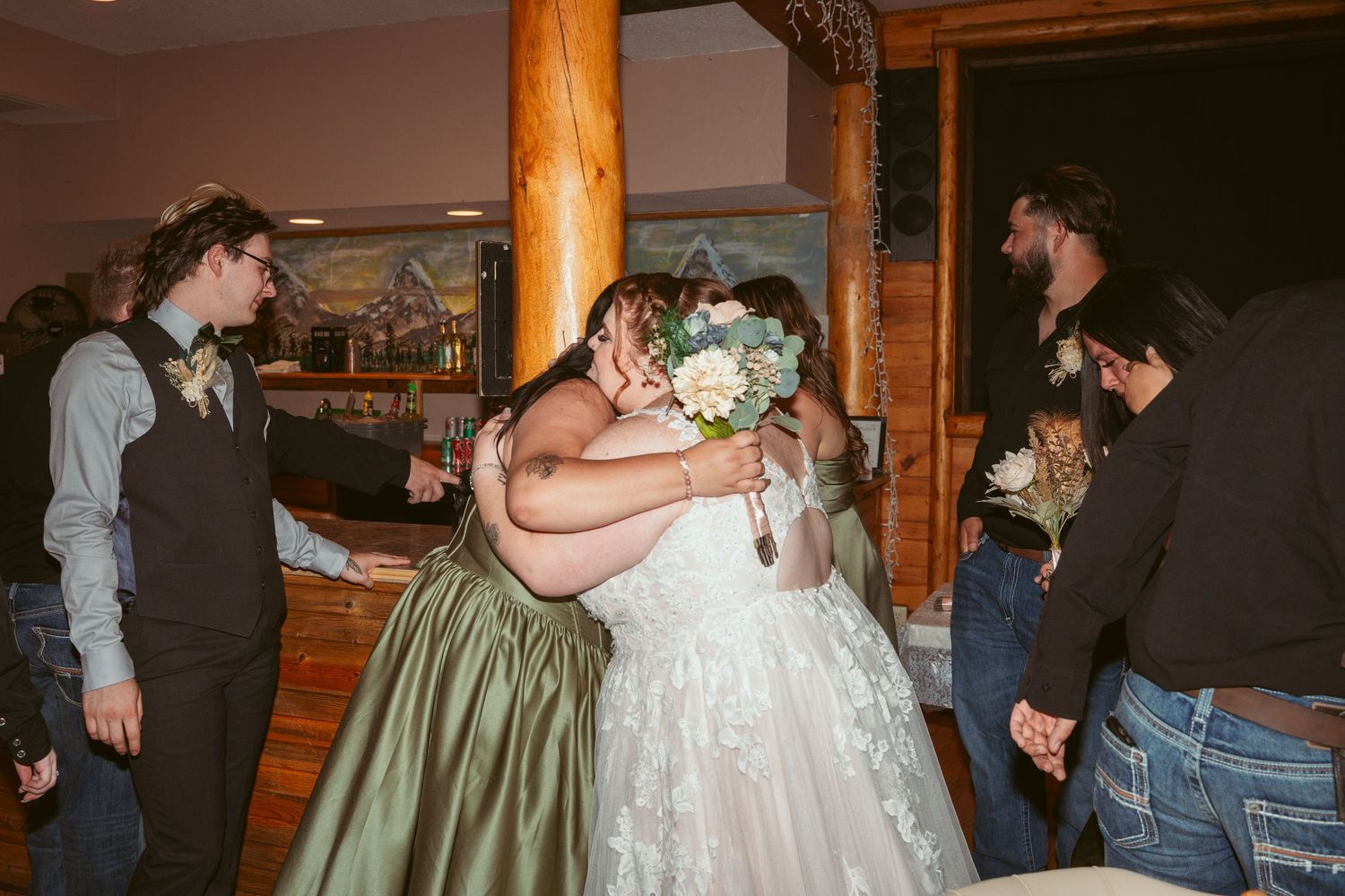 Wedding celebration scene indoors with guests gathered around a couple embracing near a wooden pillar.