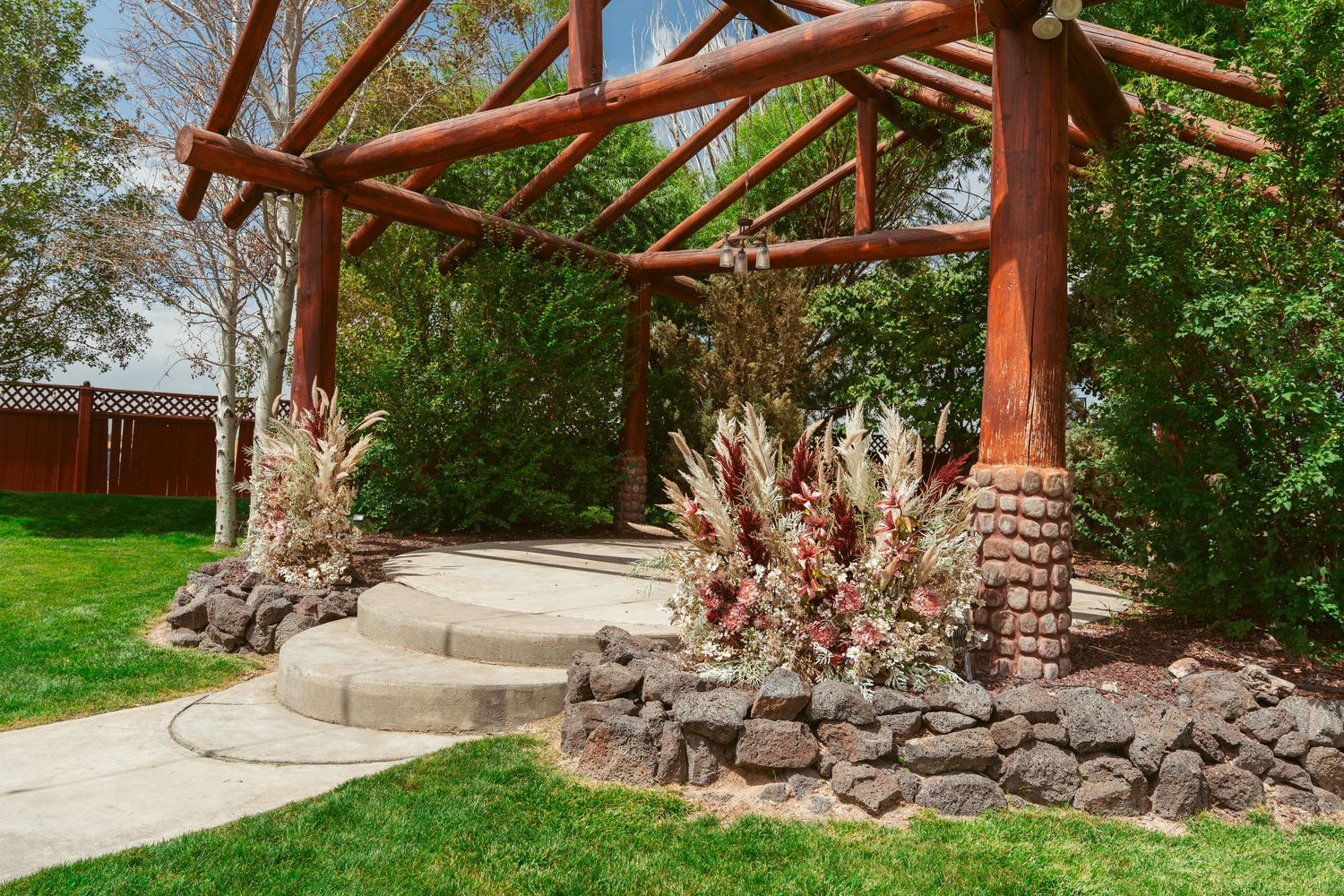 Wooden pergola structure with stone pillars along a curved concrete pathway in a garden setting.