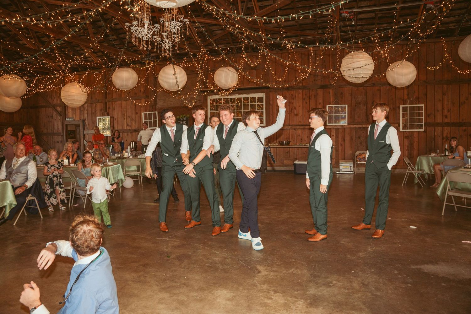 Guests dancing and celebrating at a rustic barn wedding reception with glowing paper lanterns overhead.