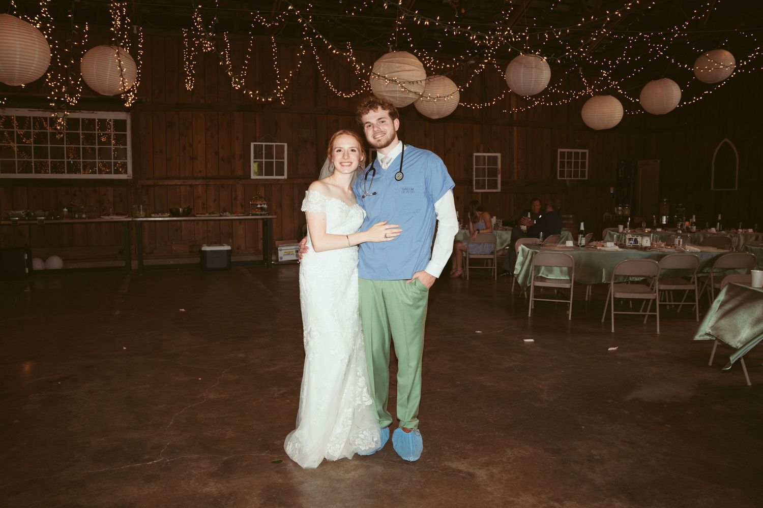 A couple dances together at a rustic barn wedding reception decorated with string lights and paper lanterns.