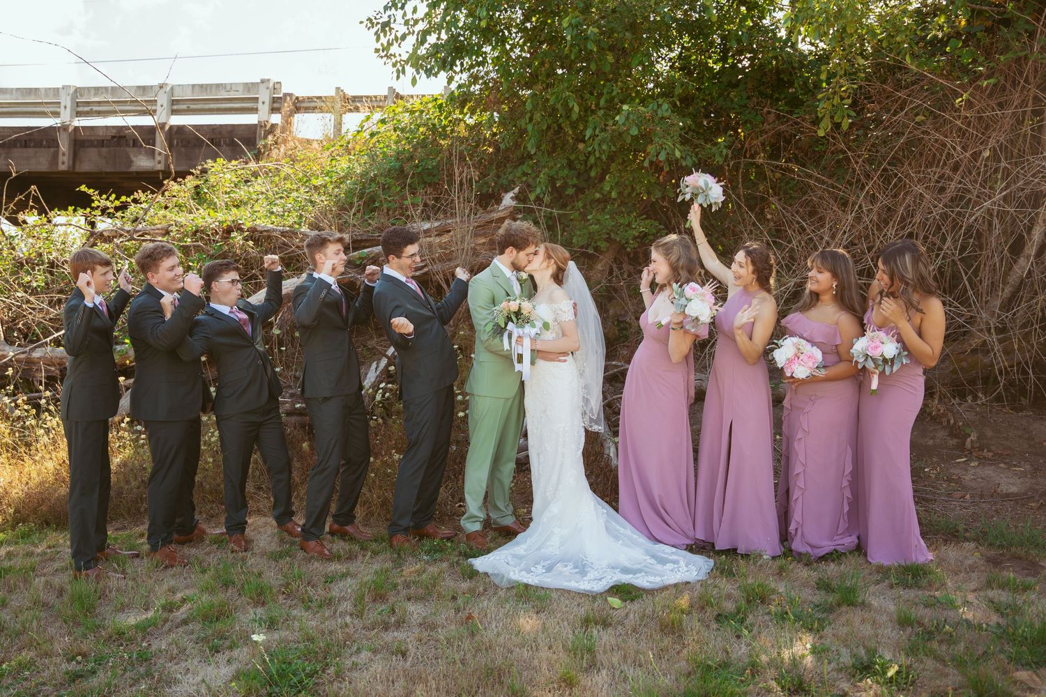 Wedding party poses together outdoors with bridesmaids in purple dresses and groomsmen in dark suits.