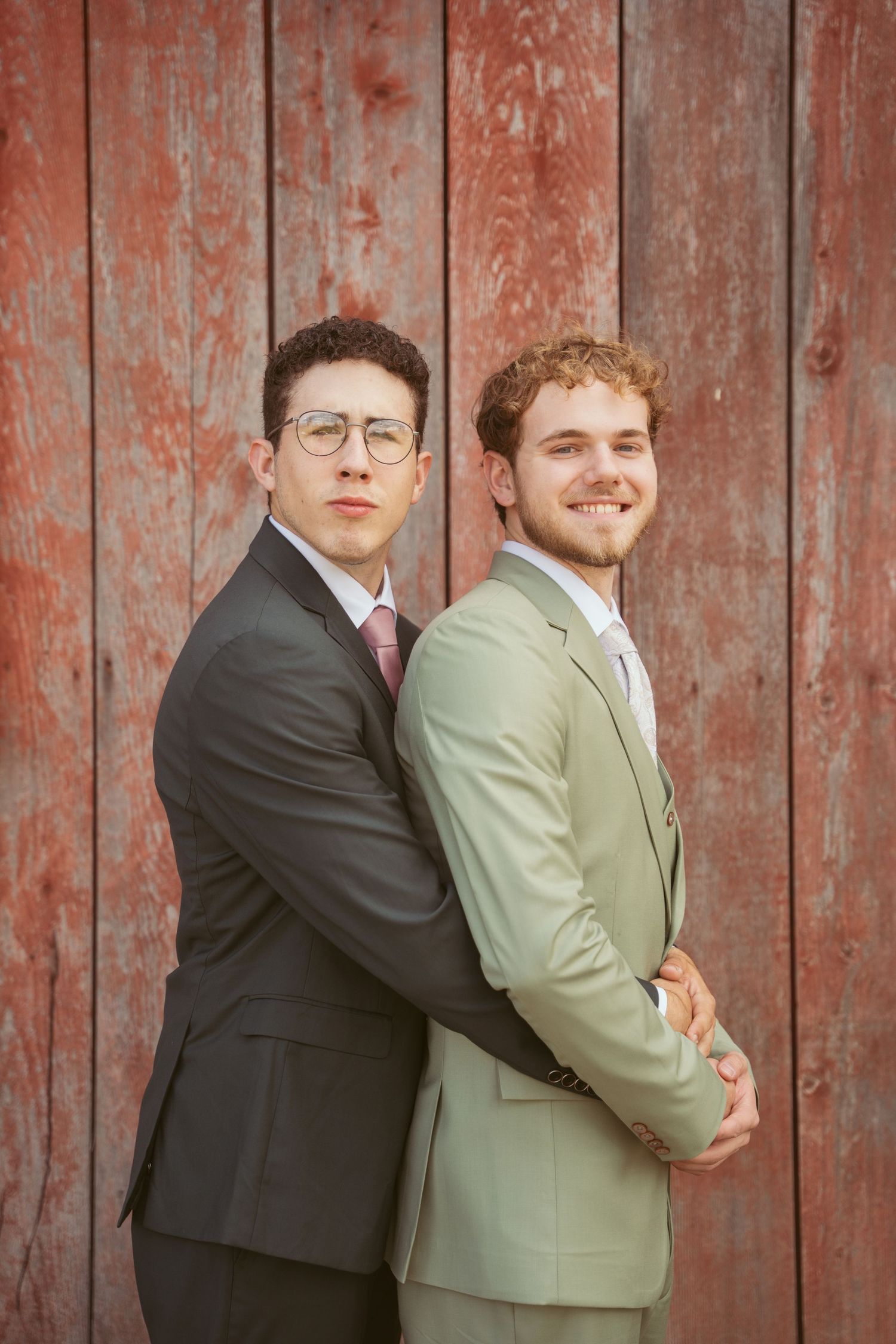 Wedding couple poses back-to-back in contrasting suits against rustic barn wall.