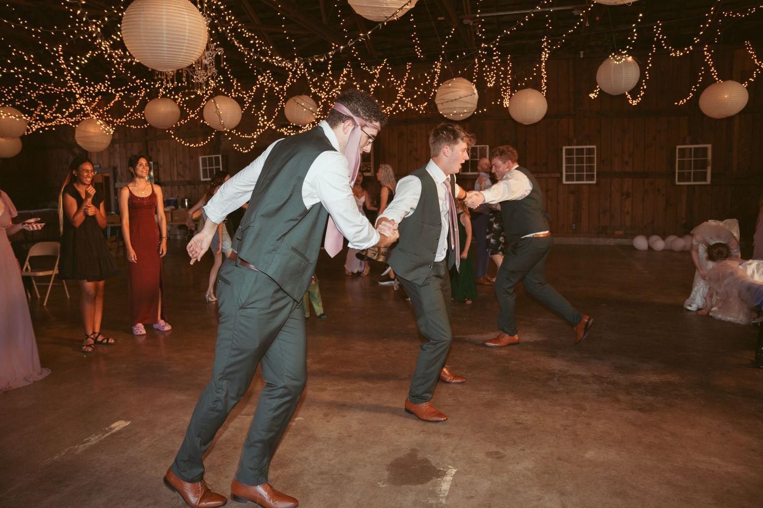 Group dance formation at a barn wedding reception under twinkling lights.