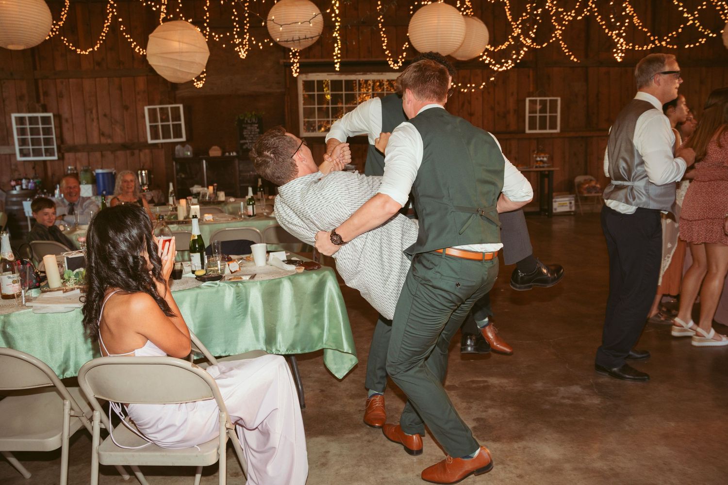 Wedding party dances beneath twinkling lights and paper lanterns at an indoor reception.