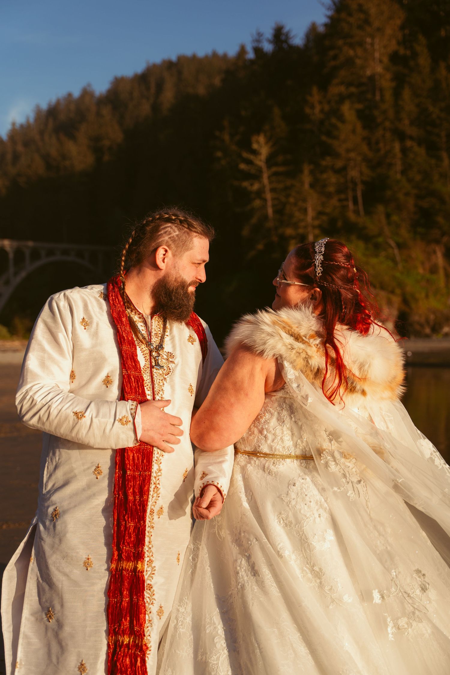 Couple in white wedding attire sharing intimate moment against forest backdrop at sunset.