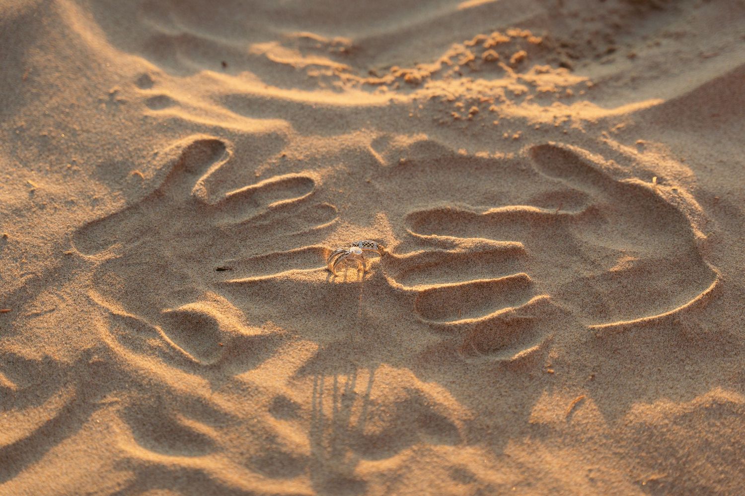 Two handprints pressed into sandy beach at sunset creating a heart shape.