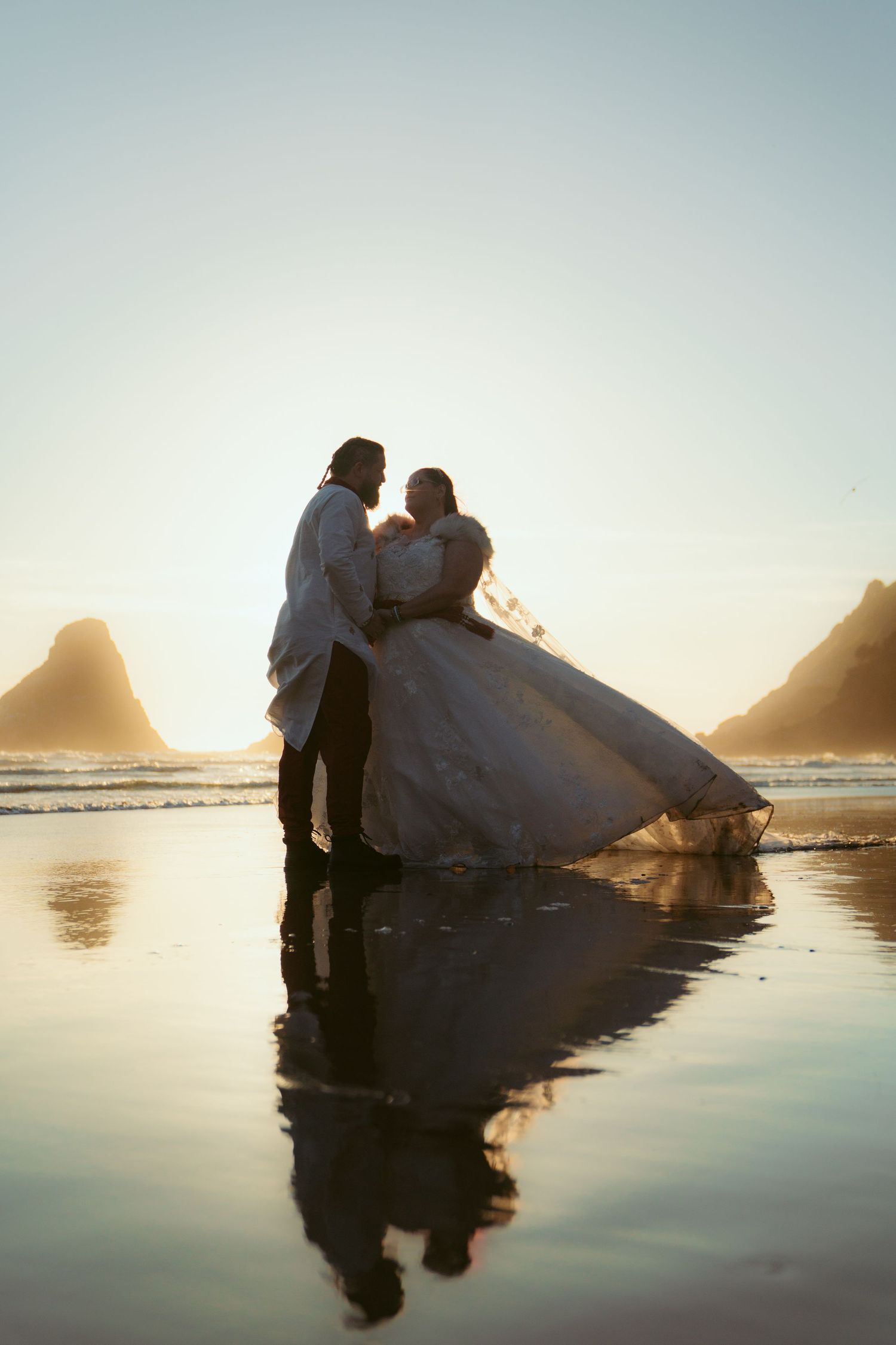 A couple in wedding attire poses for romantic photos at the water's edge during sunset.
