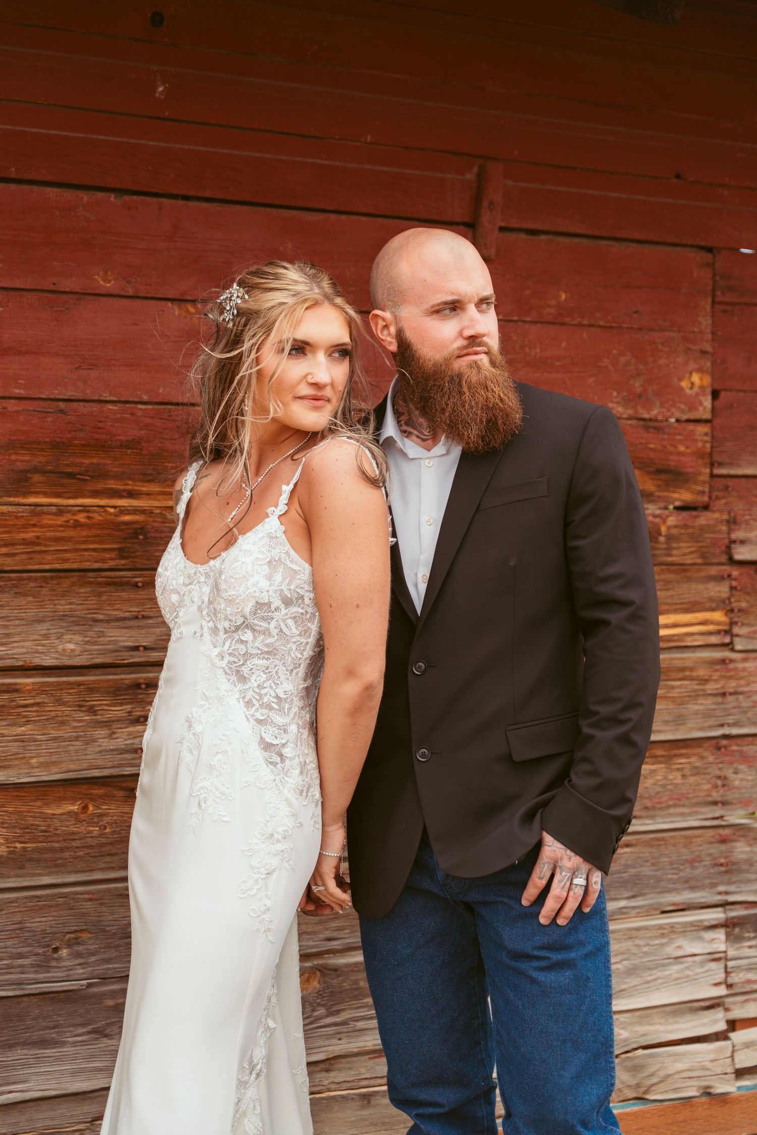 A wedding couple poses against a rustic red barn wall wearing a lace gown and casual suit.