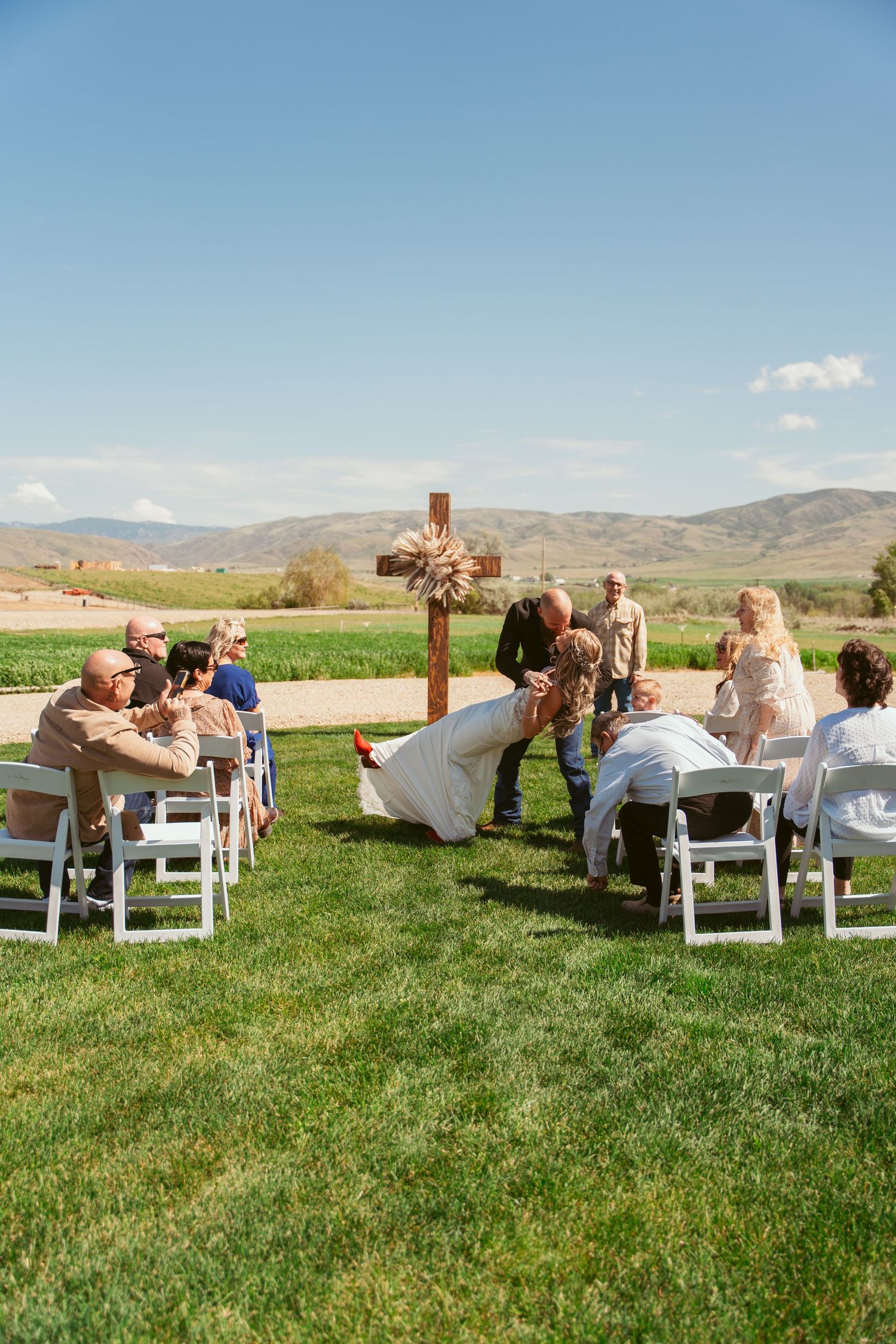 A rustic outdoor wedding ceremony with a wooden cross centerpiece and mountain landscape backdrop.
