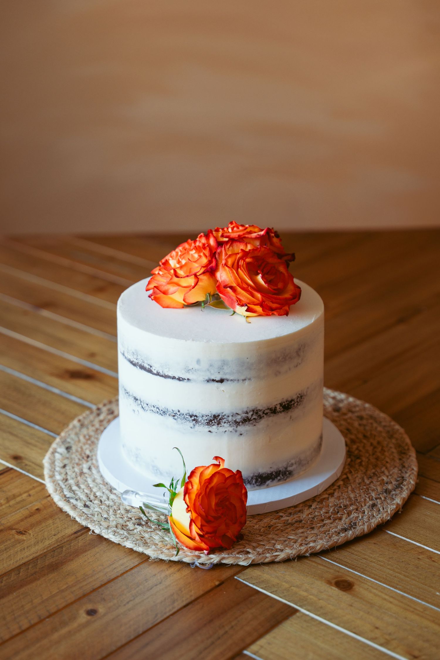 White wedding cake decorated with vibrant orange roses on a woven placemat against wooden surface.