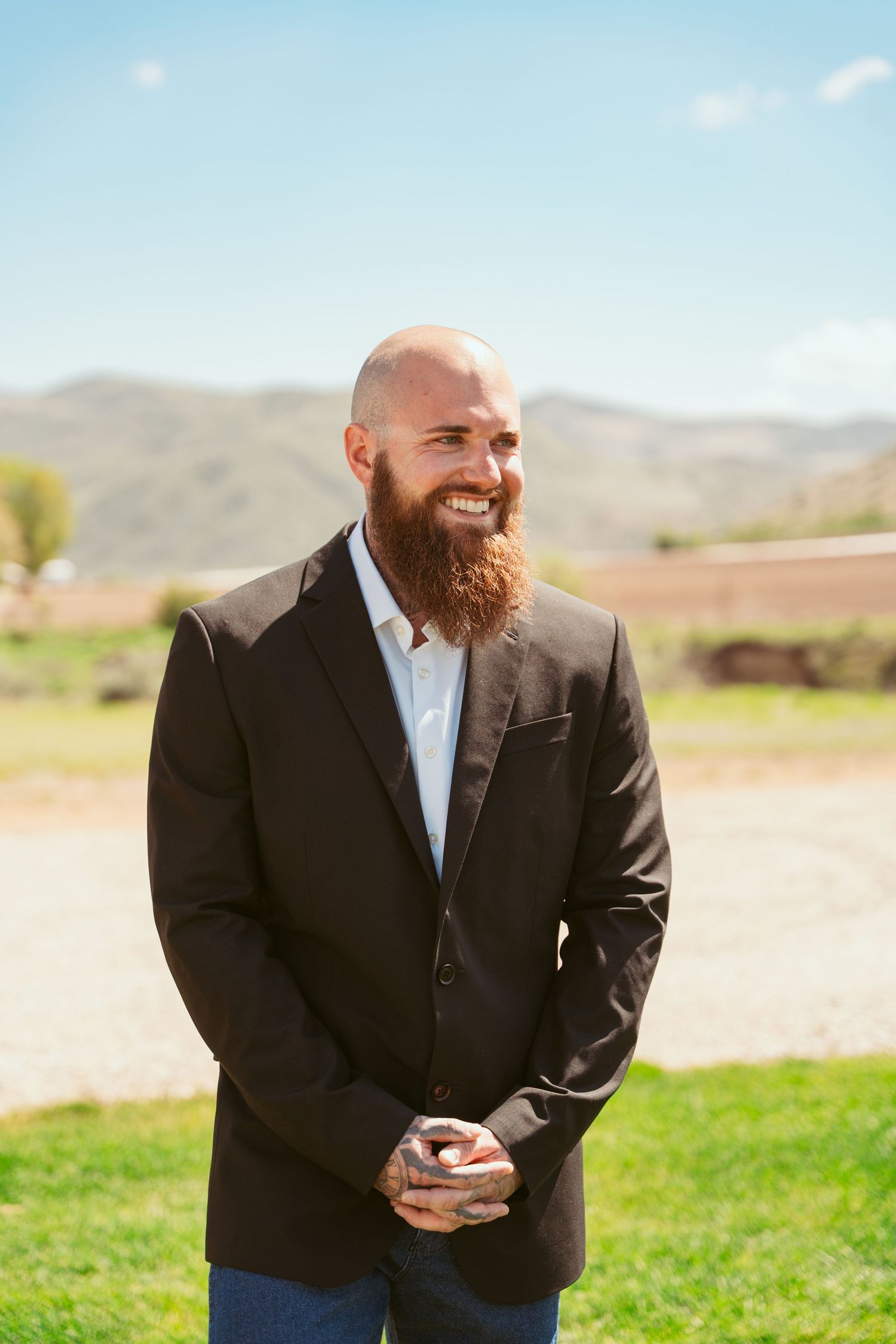 A groom in a black suit stands outdoors against a mountain landscape.