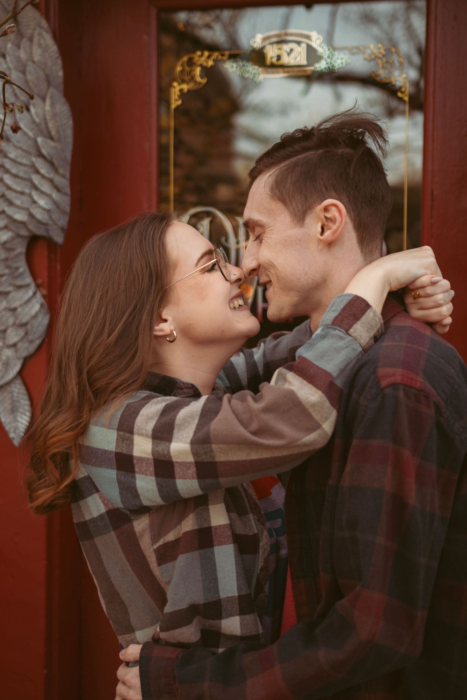 A romantic couple in plaid shirts embracing near a red doorway.