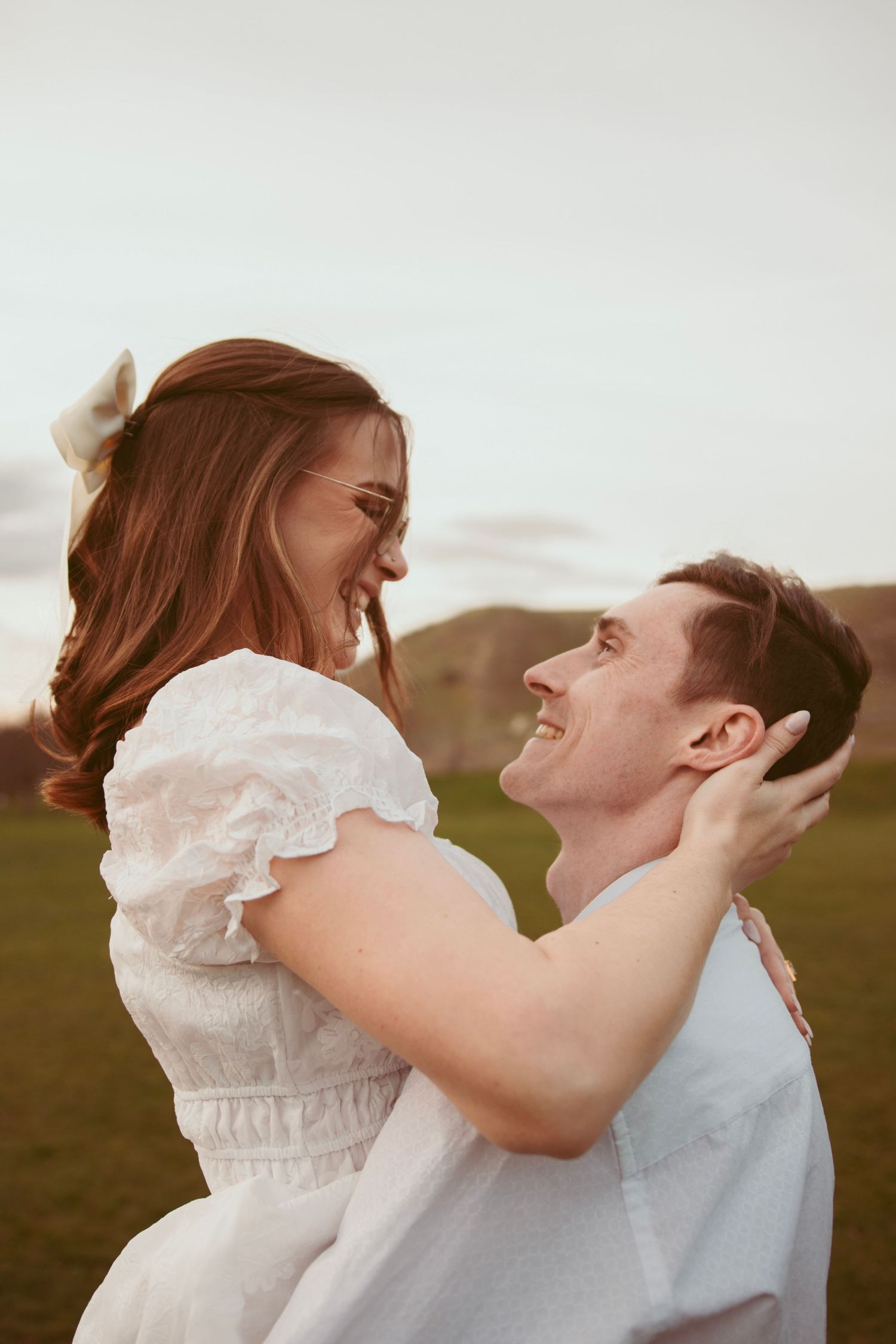 A romantic couple in white clothing embraces playfully in a grassy field during golden hour.