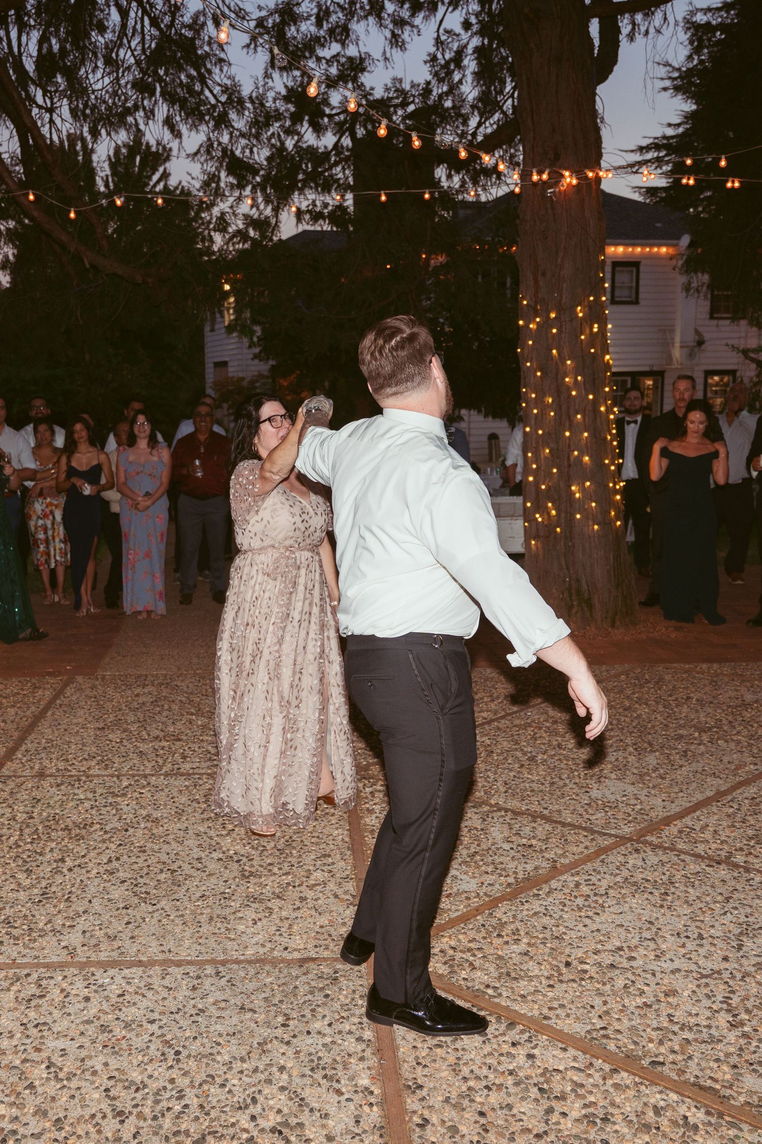Evening wedding reception with guests dancing on gravel under string lights and trees.