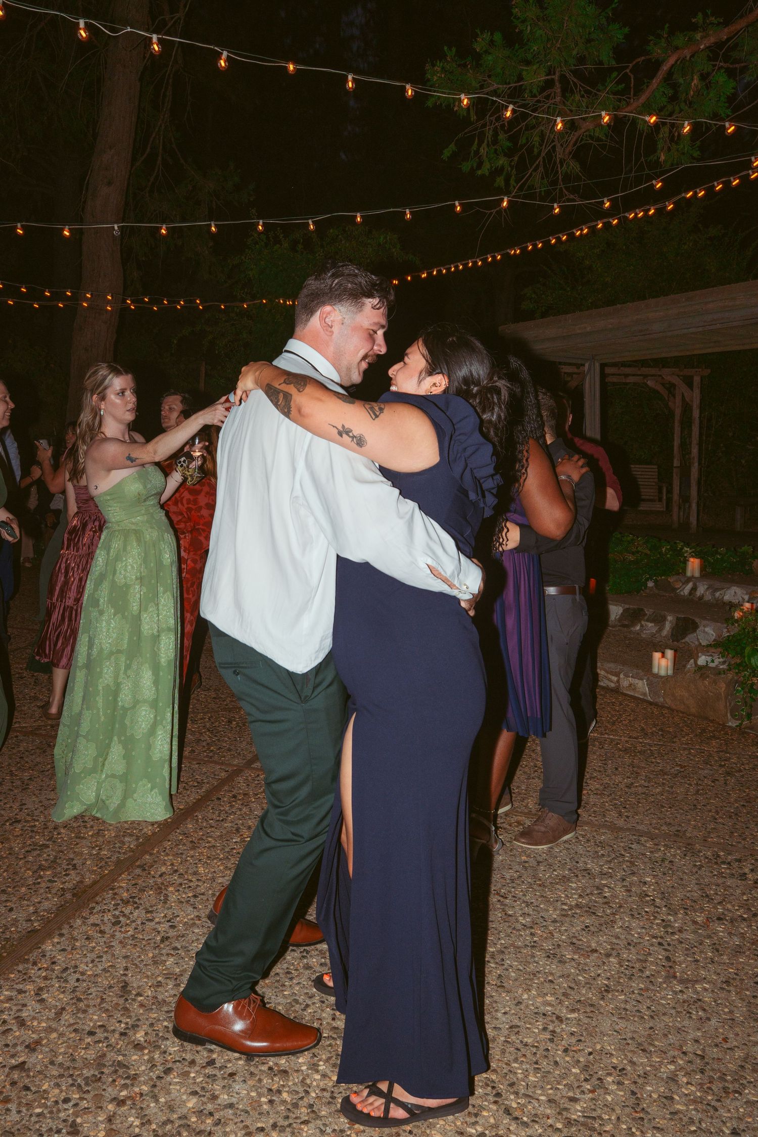 People dancing together under string lights at an outdoor evening celebration with a rustic setting.