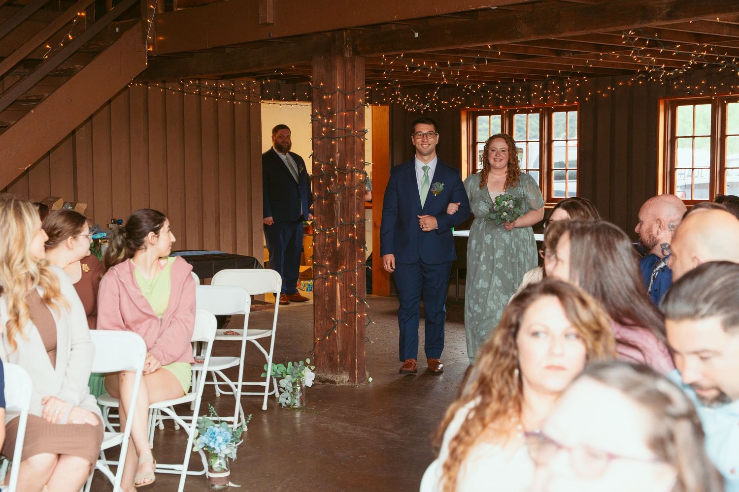 Guests enjoying a celebration in a wooden barn with twinkling lights and folding chairs.