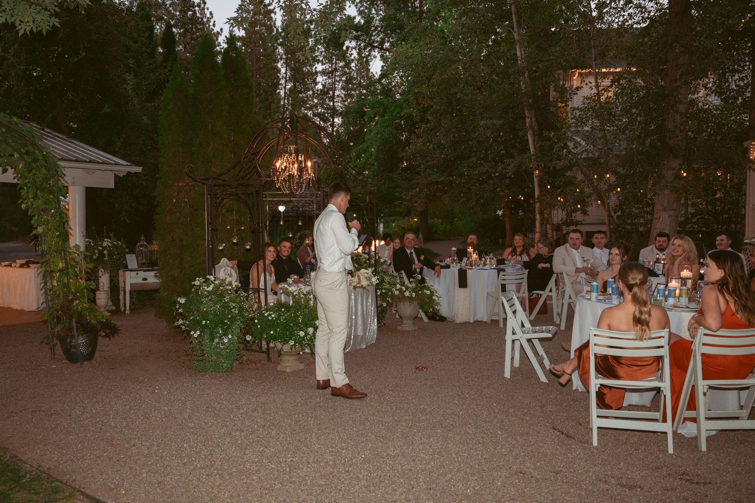 Evening outdoor wedding reception with white chairs and tables set among trees and string lights.