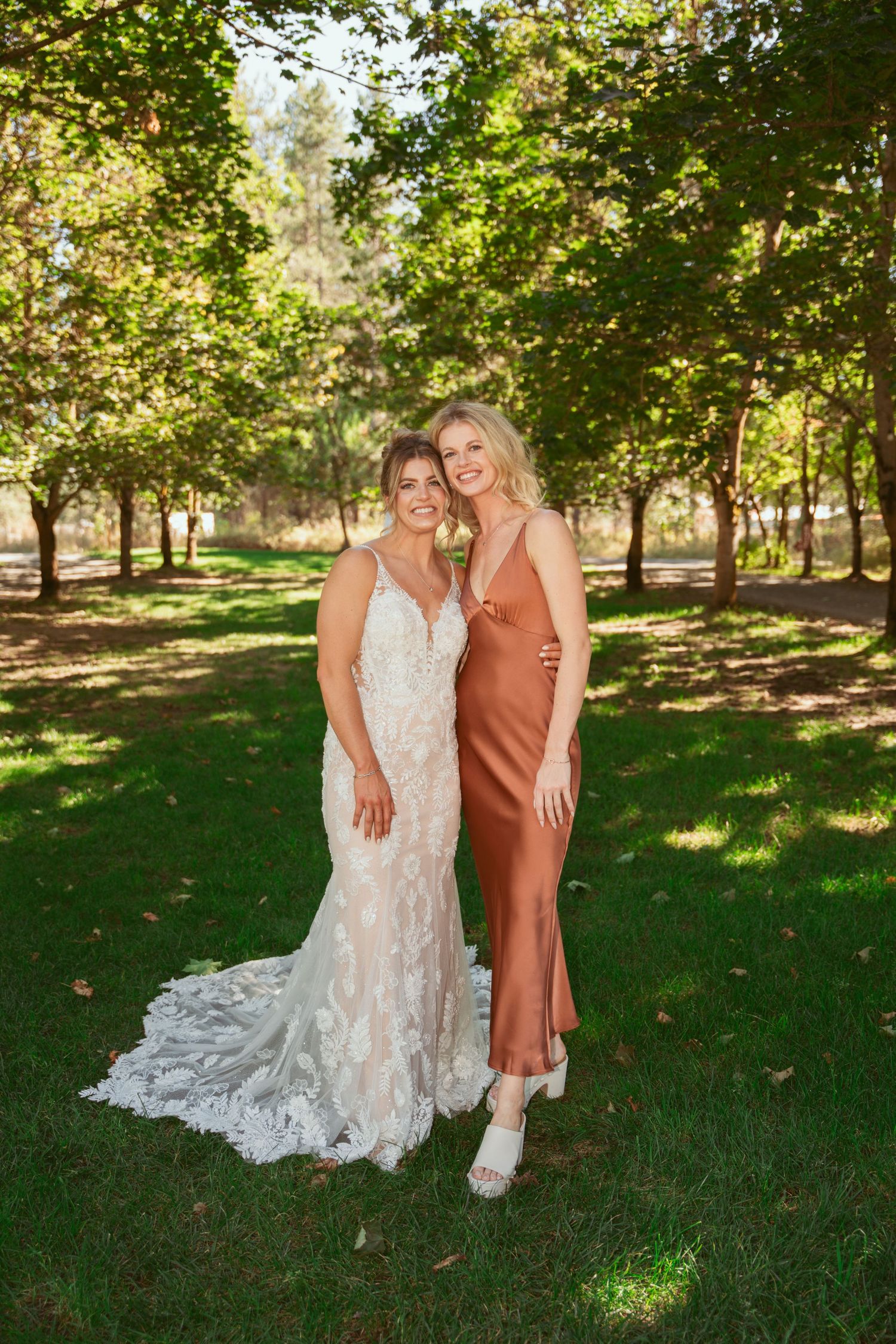 Two people pose together outdoors in a park setting with sunlit trees, one wearing a lace wedding gown and another in silk dress.