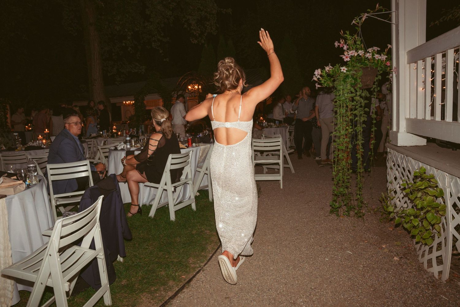 A wedding guest in a white dress dances joyfully between rows of outdoor reception tables at dusk.