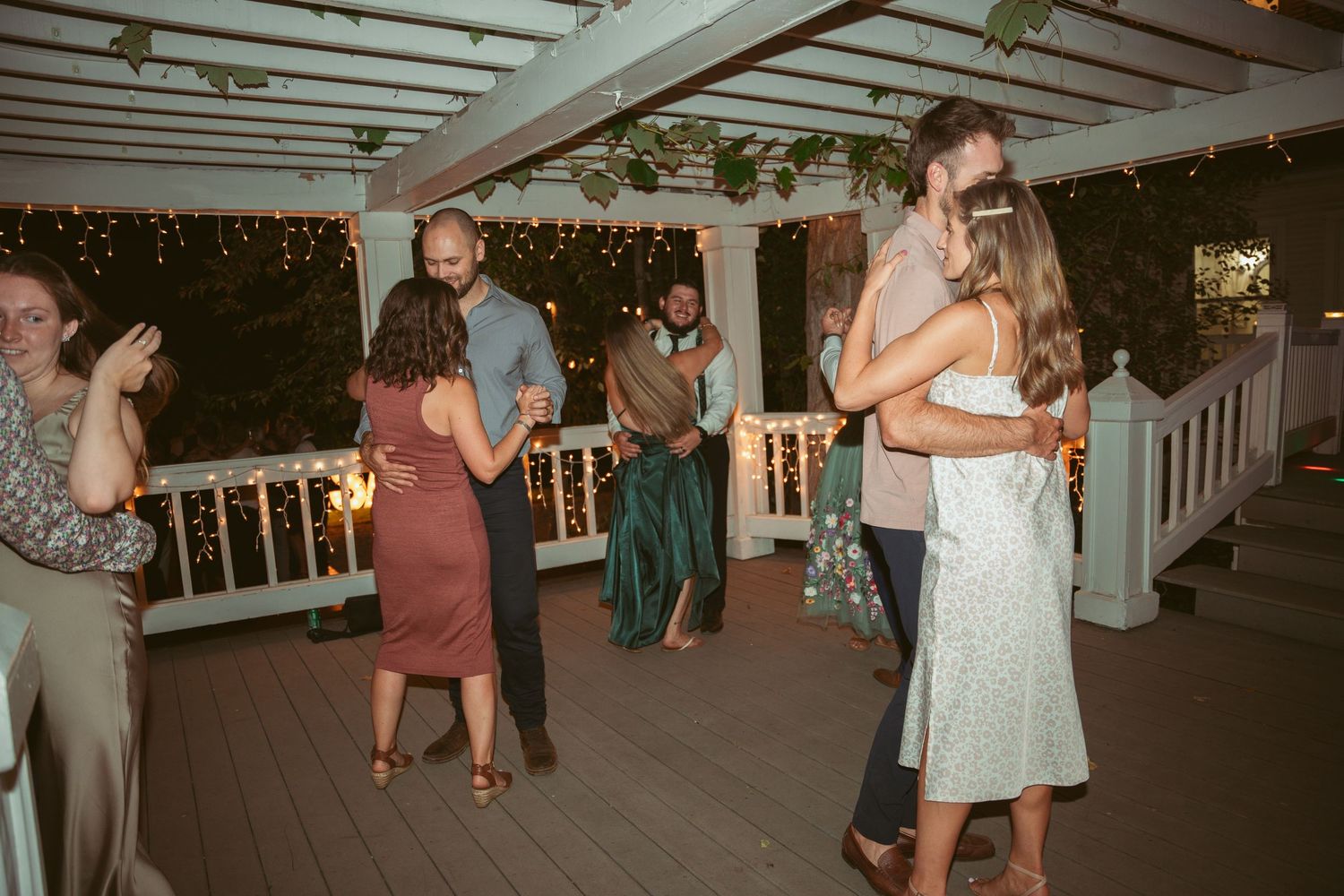 People dancing and celebrating at an outdoor evening party under a white pergola with string lights.