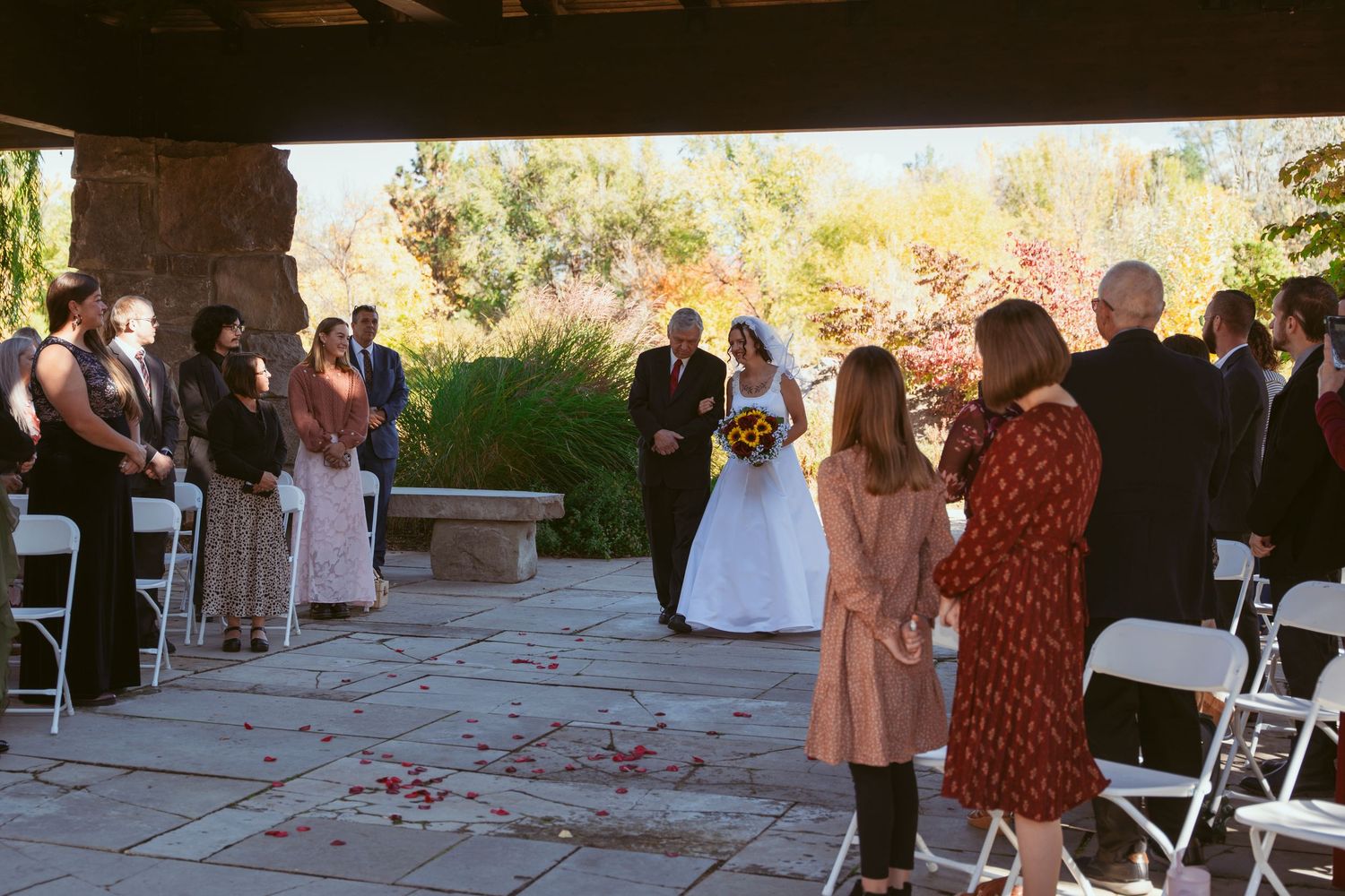 Wedding ceremony taking place outdoors on a stone patio with scattered rose petals and white chairs.