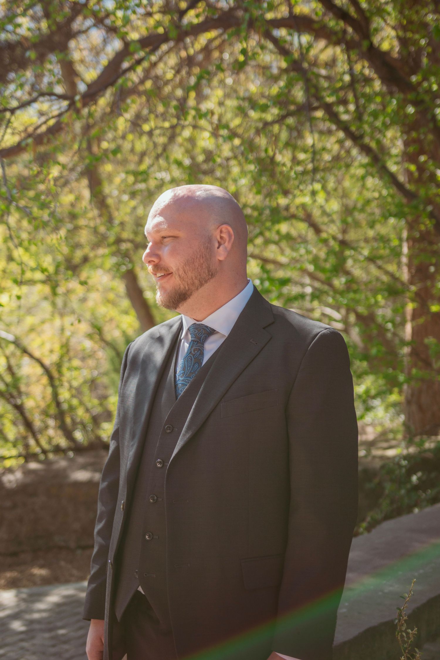 A groom in a dark suit and blue tie stands in dappled sunlight beneath trees during an outdoor wedding.