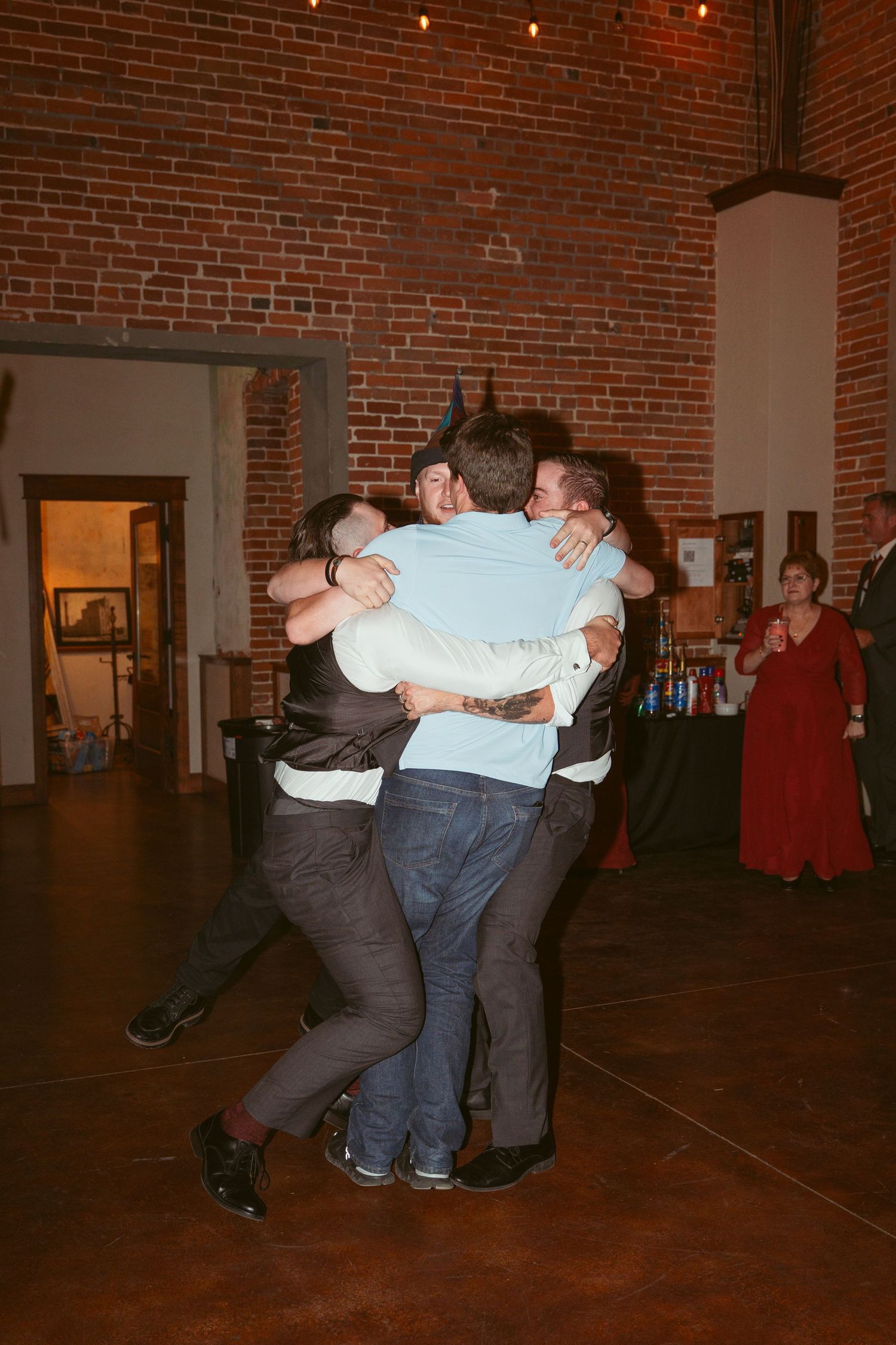 Friends sharing a group hug on a dance floor in a rustic brick venue with string lights.