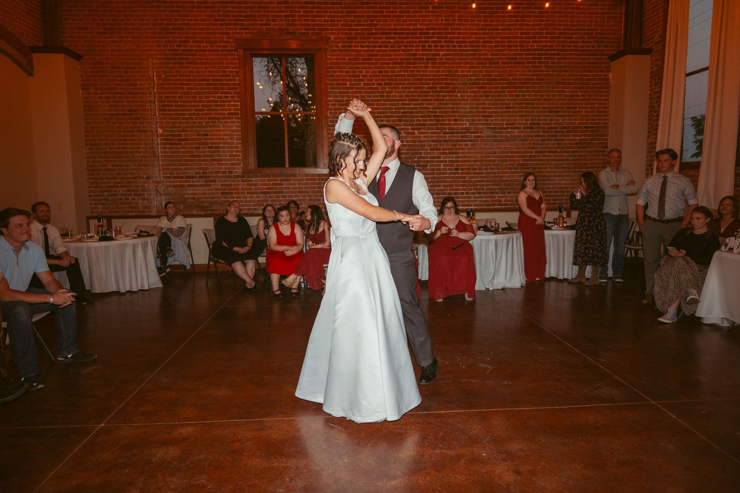 A couple shares their first dance in a rustic brick-walled venue with string lights overhead.