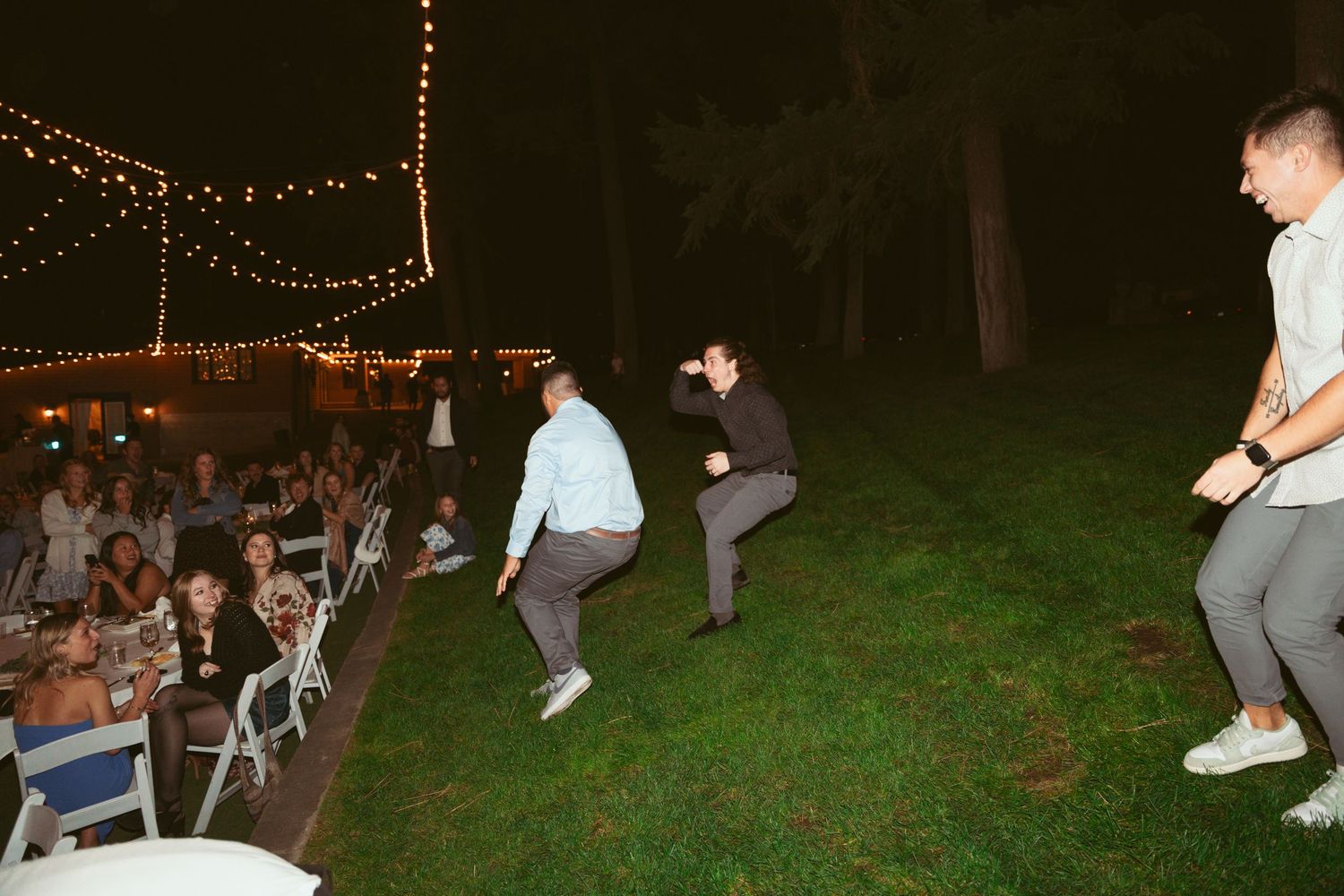 String lights illuminate an outdoor evening party scene with people dancing and mingling on a grassy lawn.
