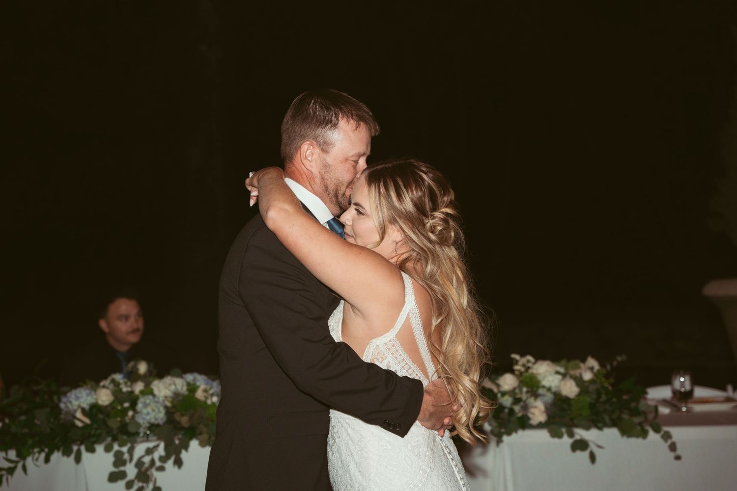 A couple shares their first dance at a wedding reception with white floral decorations in the background.