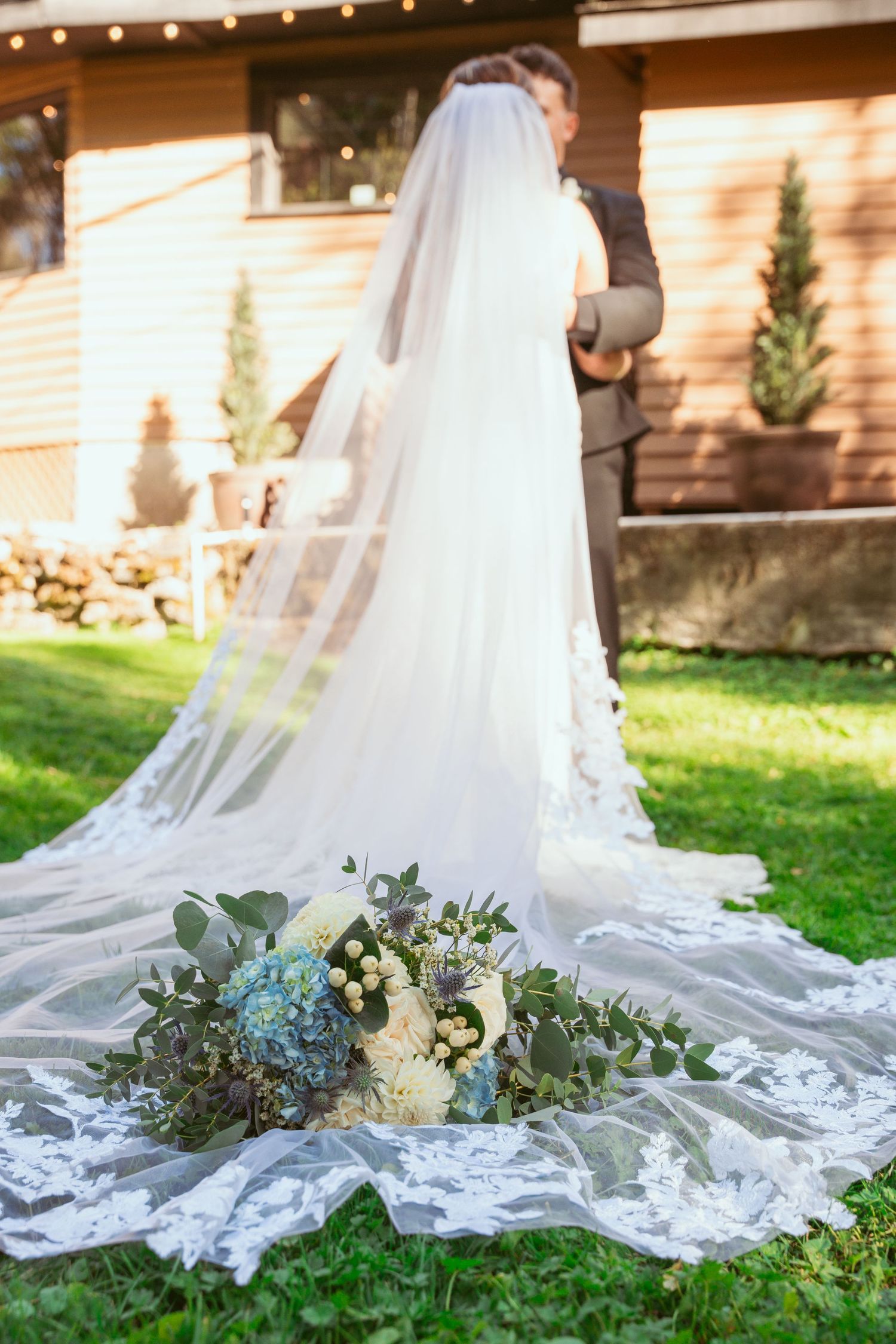 Elegant white wedding dress train with delicate lace details and floral bouquet arranged on green grass.