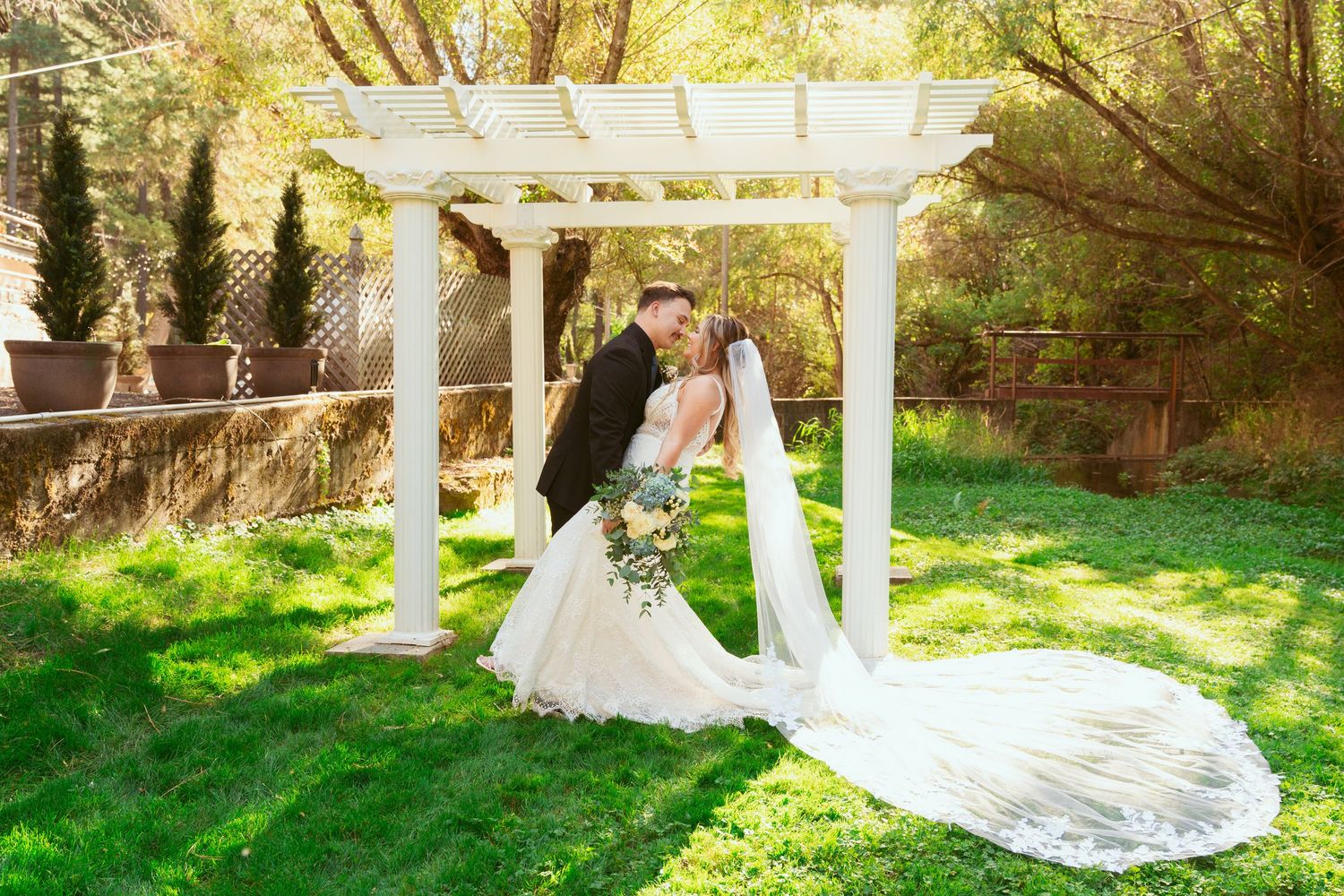 Wedding party gathers for photos under an elegant white pergola in a sunlit garden setting.