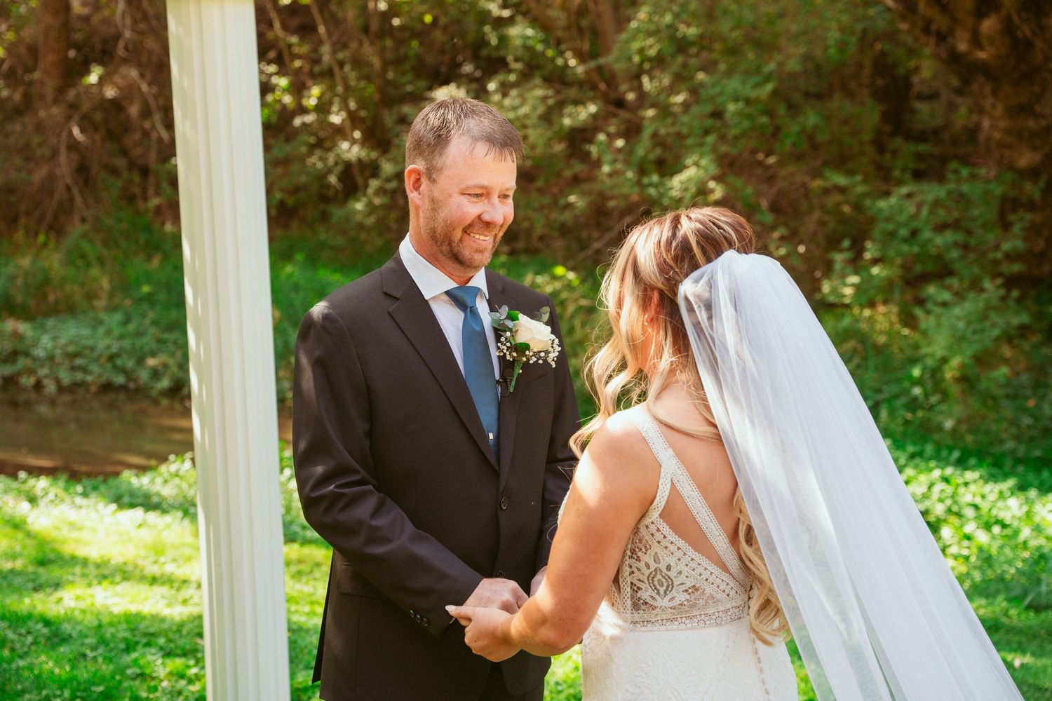 A couple shares a romantic moment during their outdoor wedding ceremony beneath white pillars and lush greenery.