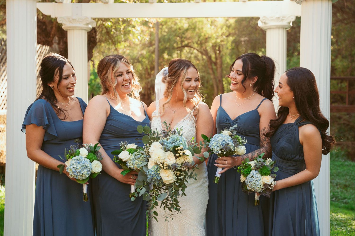 Bridesmaids in blue dresses share candid moment with bride under white pergola.
