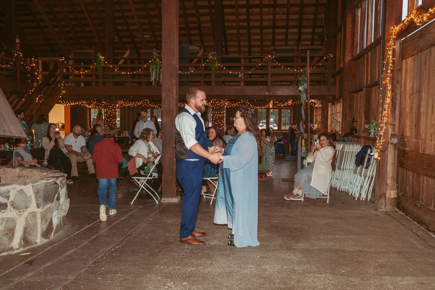 Wedding guests dance and celebrate in a wooden barn decorated with twinkling lights.